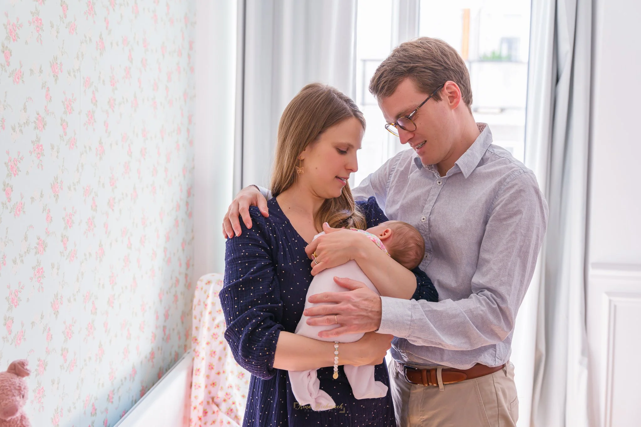 Nouvelle maman et nouveau papa regardent leur bébé nouveau né dormir dans les bras de sa maman dans une chambre baignée de lumière naturelle 
