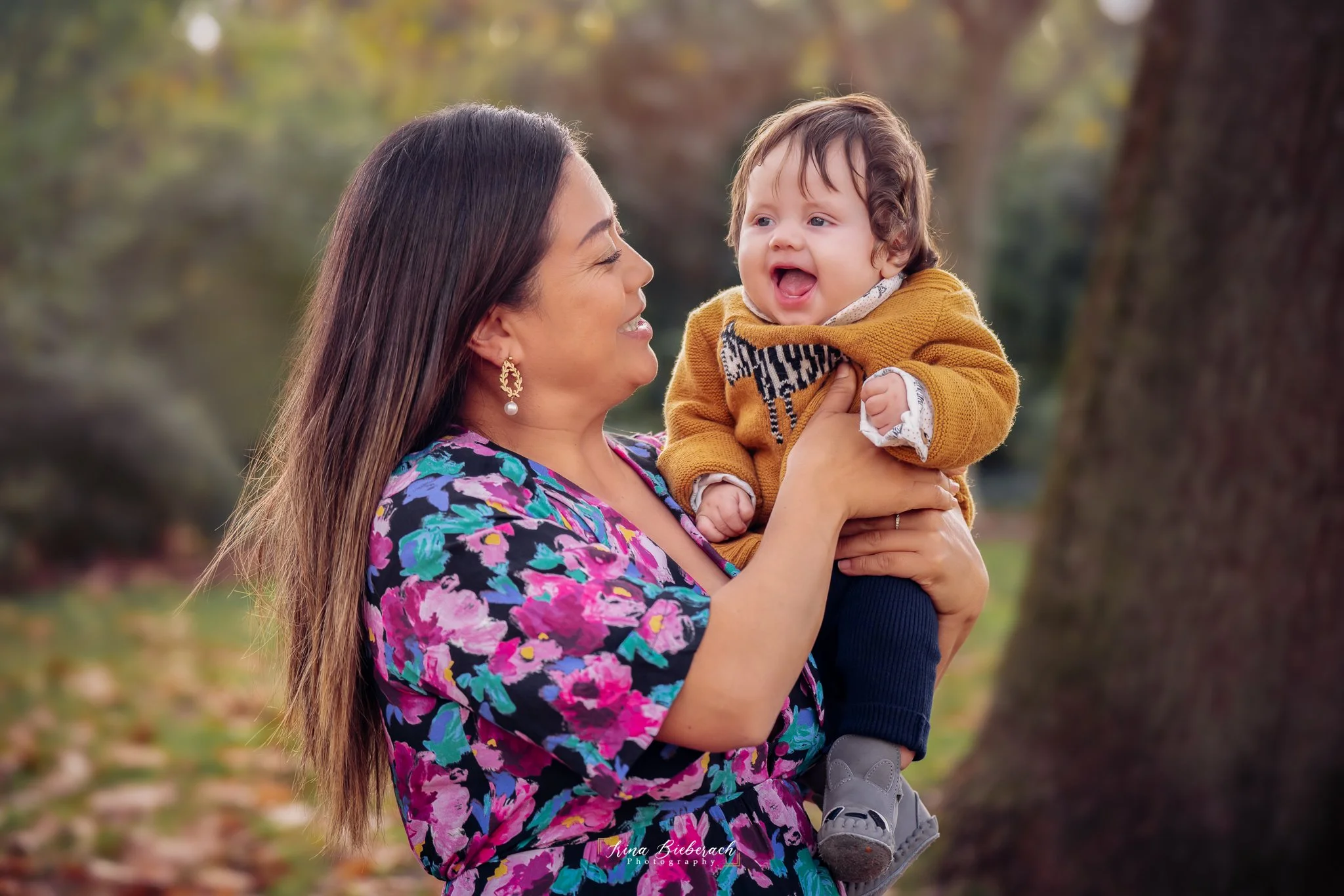 Femme brune porte son bébé souriant et le regarde rire dans  le parc Montsouris 