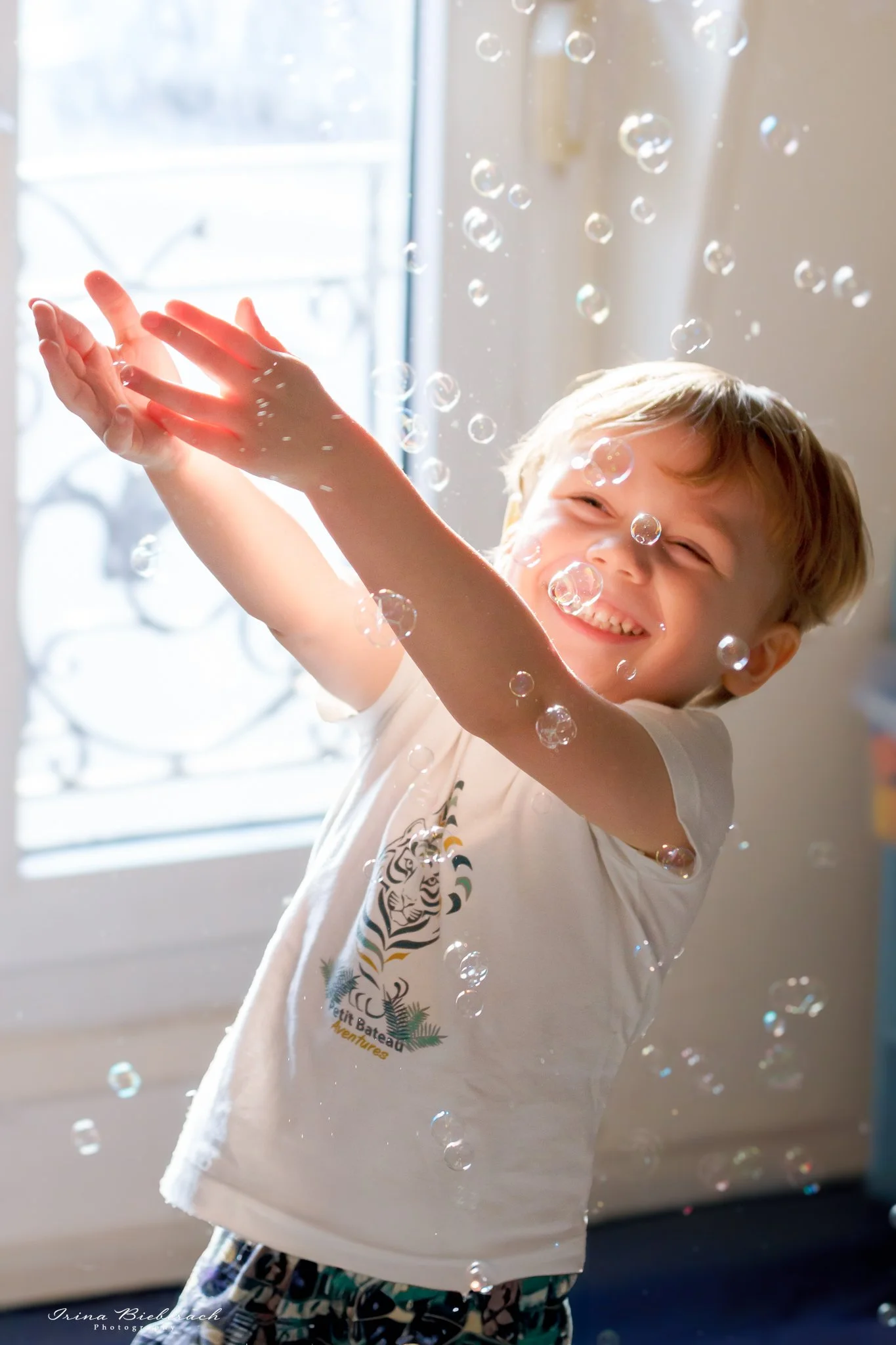 Petit garçon souriant essaye d’attraper les bulles de savons dans ses mains dans une chambre d’appartement parisien