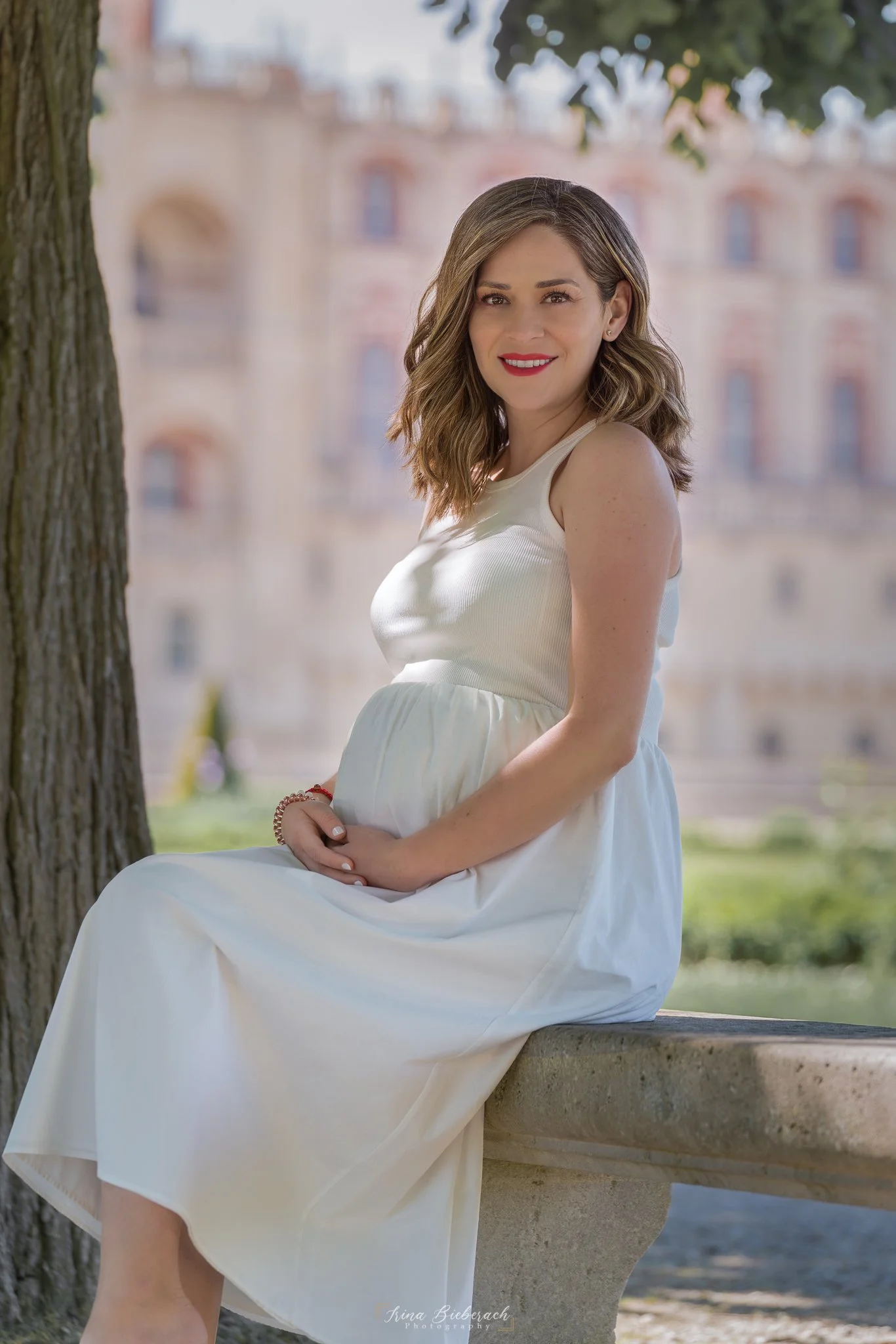 Futur maman souriante et habillée en robe blanche pose assise devant le château de Saint Germain en Laye