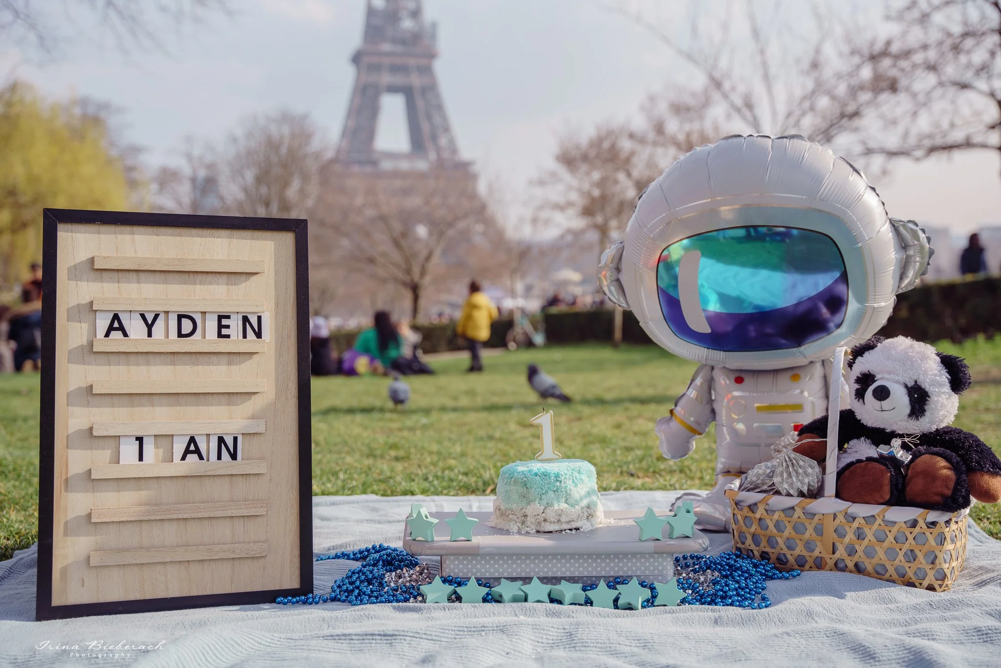 Décoration pour  Smash the cake devant la Tour Eiffel avec  gâteau bleu pastel, nappe de pique nique, ballon astronaute, peluche ourson dans un panier et tableau de prénom 