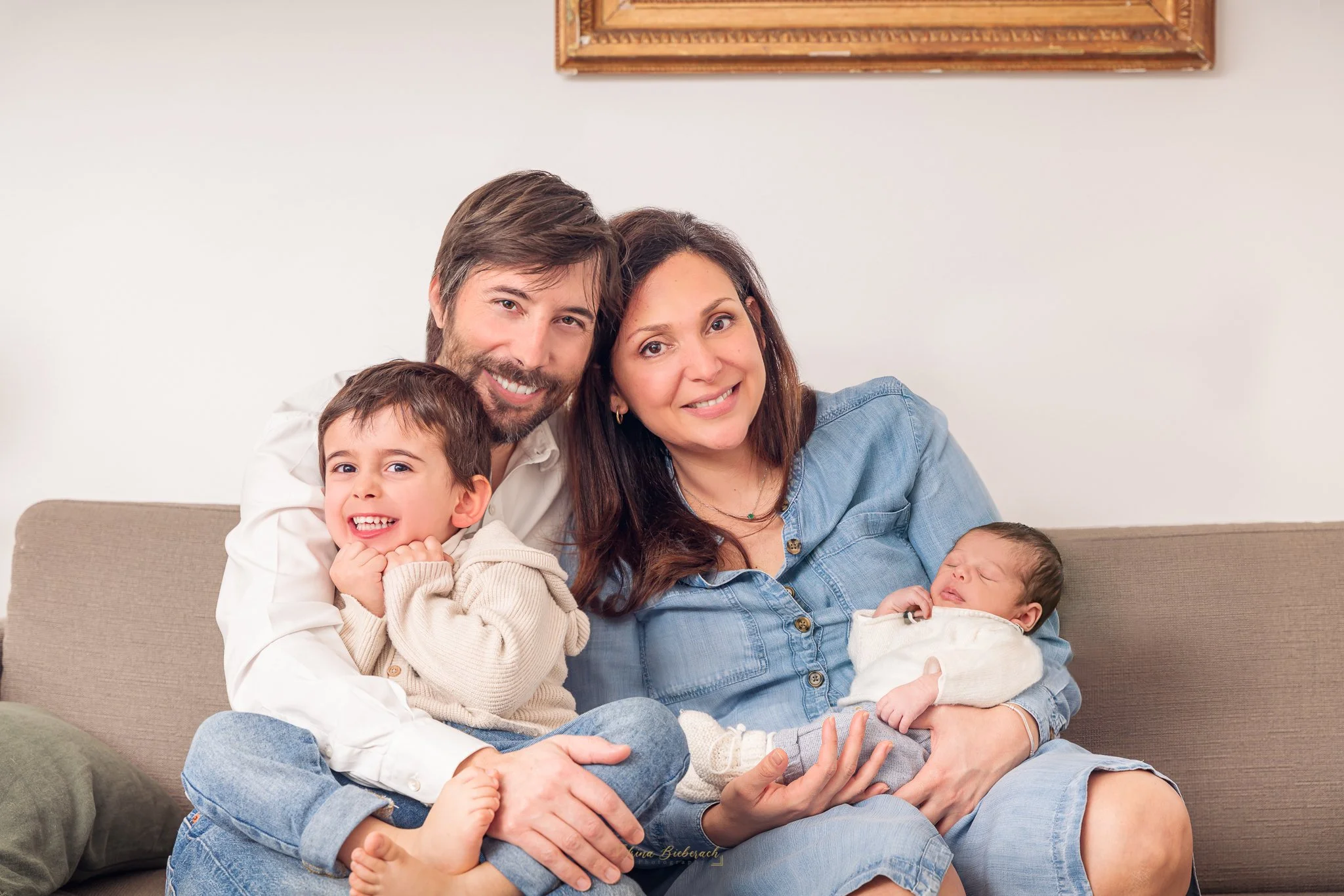 Portrait d'une famille avec maman qui porte le nourrisson dans ses bras et papa qui porte le fils ainé sur ses genoux en souriant dans un canapé de salon