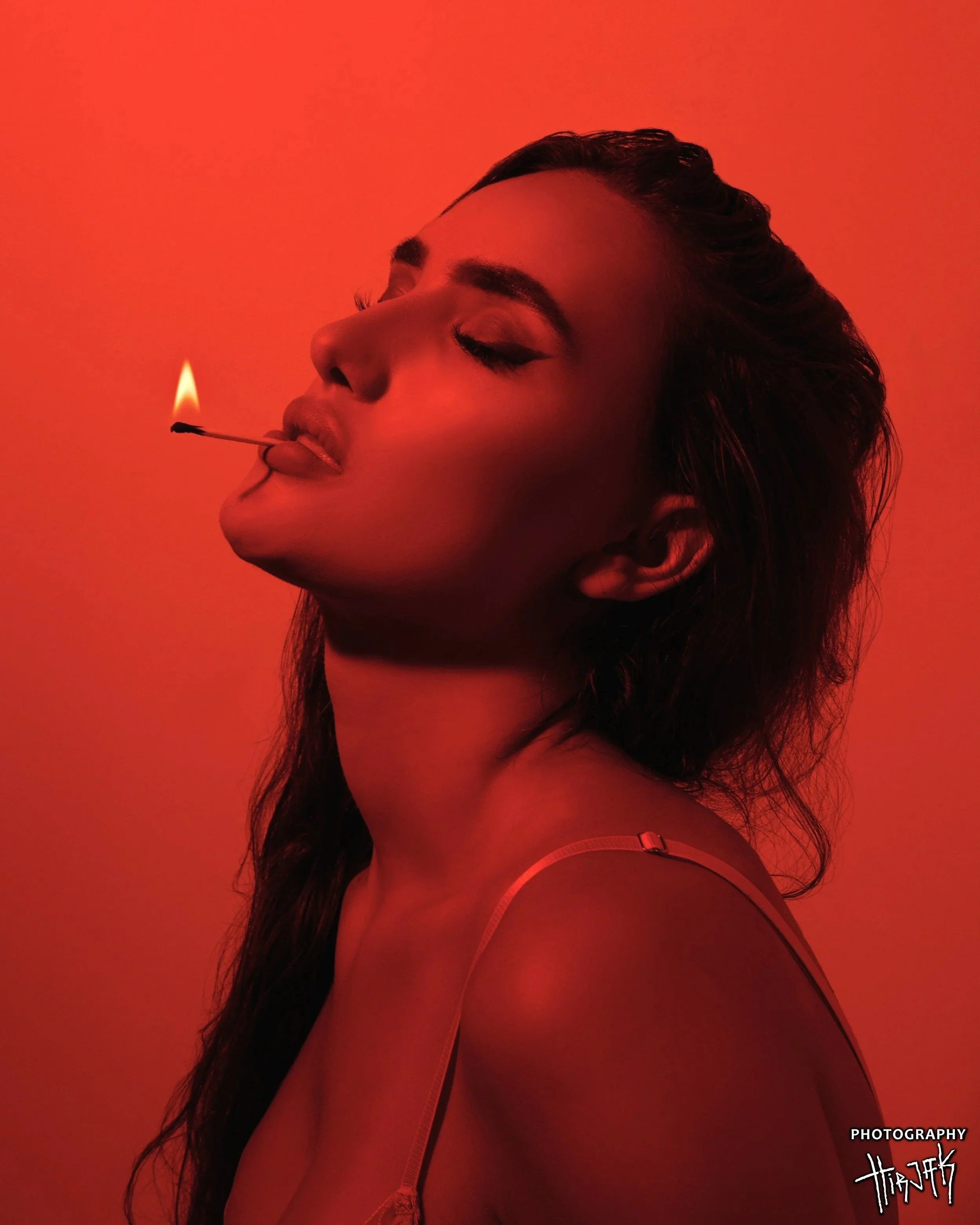 A woman with closed eyes and long dark hair holding a lit matchstick in her mouth against a red background.