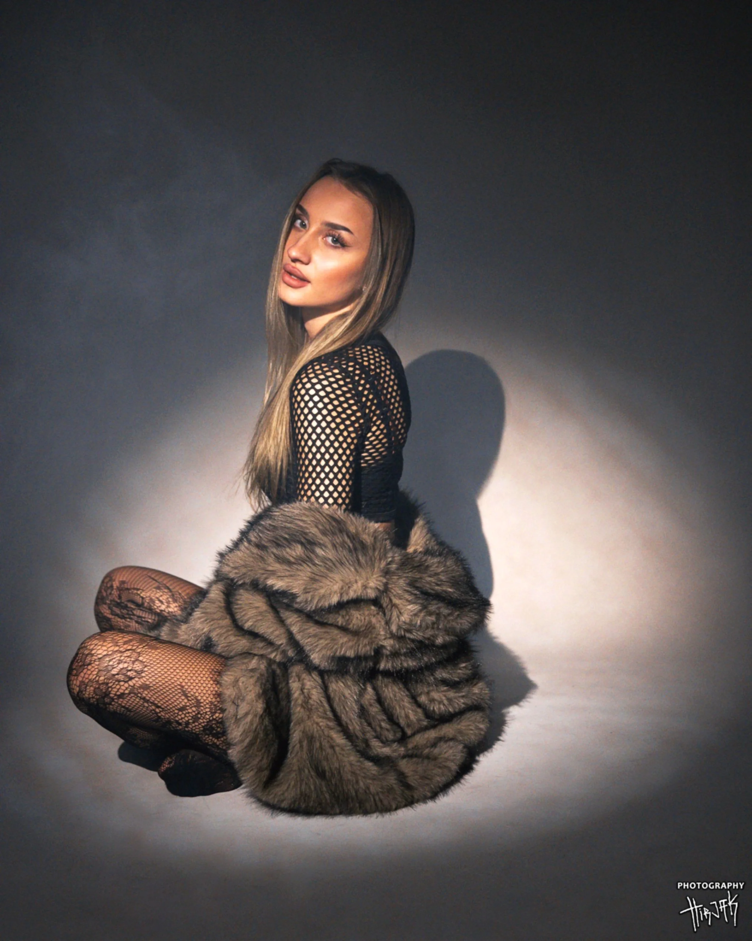 A woman with long, brown hair and light makeup sitting on the floor against a dark background. She's wearing a black fishnet top and patterned tights, and holding a large fur coat. She gazes at the camera with a confident expression.