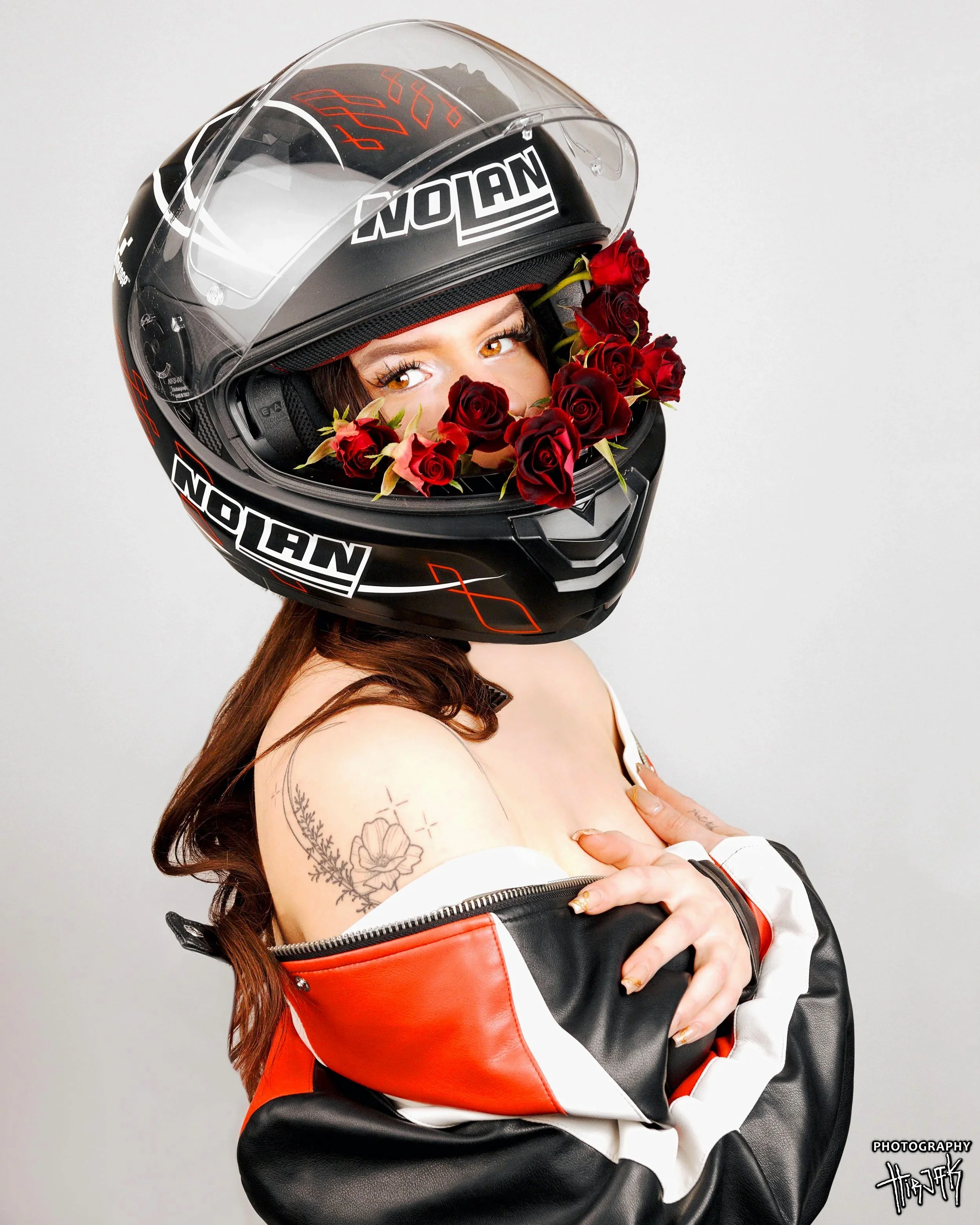 A woman wearing a motorcycle helmet decorated with red roses, looking through the helmet's visor. She has tattoos on her arm, and is dressed in a black and red leather jacket. The background is plain white.