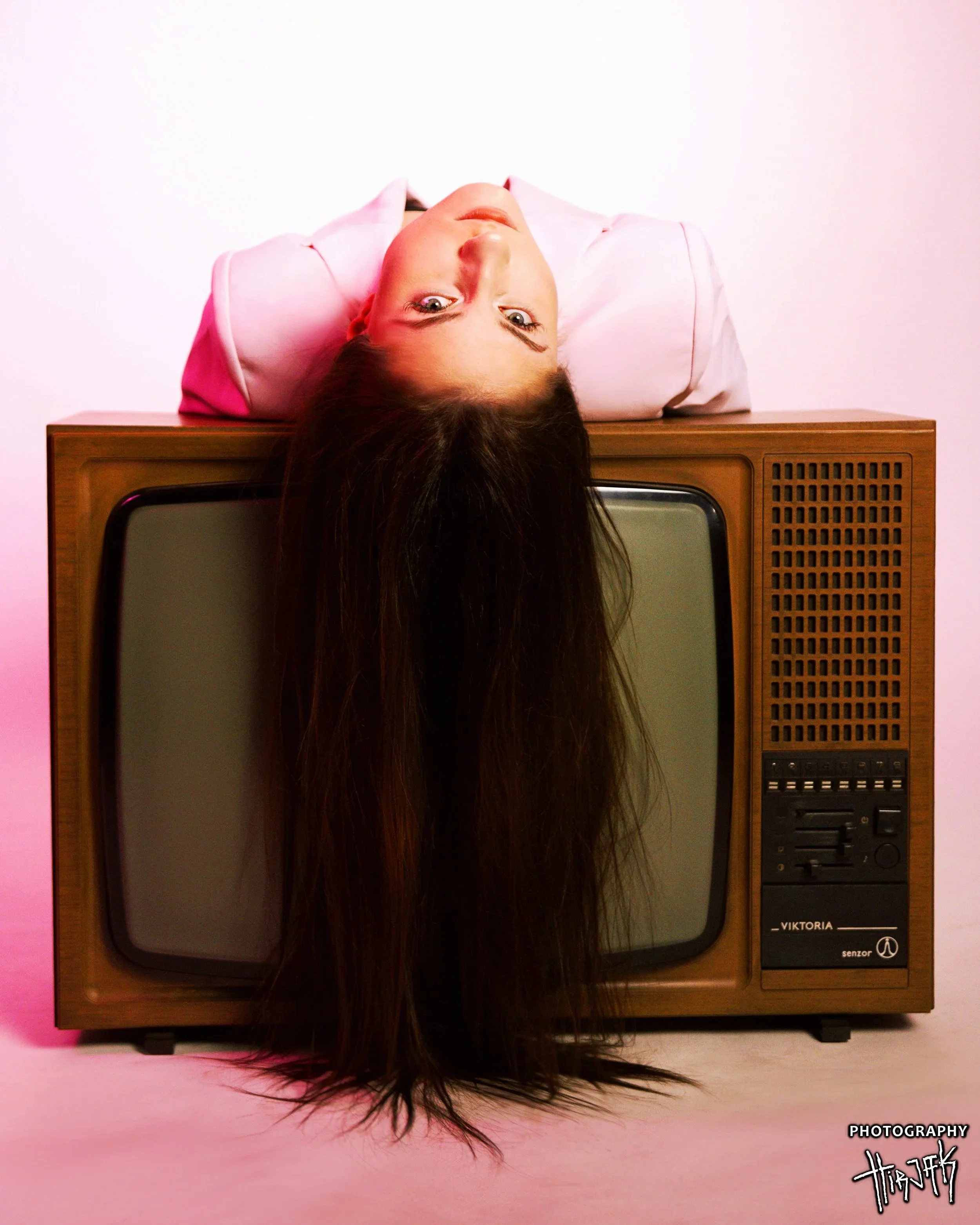 A woman with long dark hair lying on top of an old wooden television set, facing upward with her head hanging over the edge.