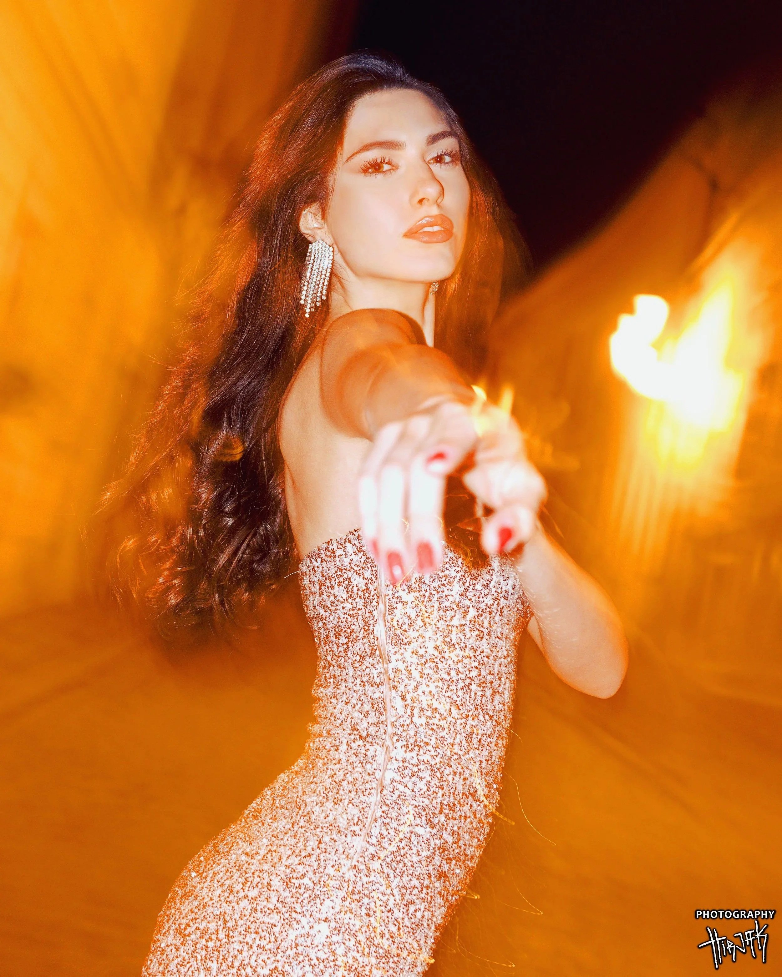 A woman with long dark hair, wearing sparkling earrings and a glittering dress, points a finger towards the camera in a nighttime setting with warm orange and yellow lights.