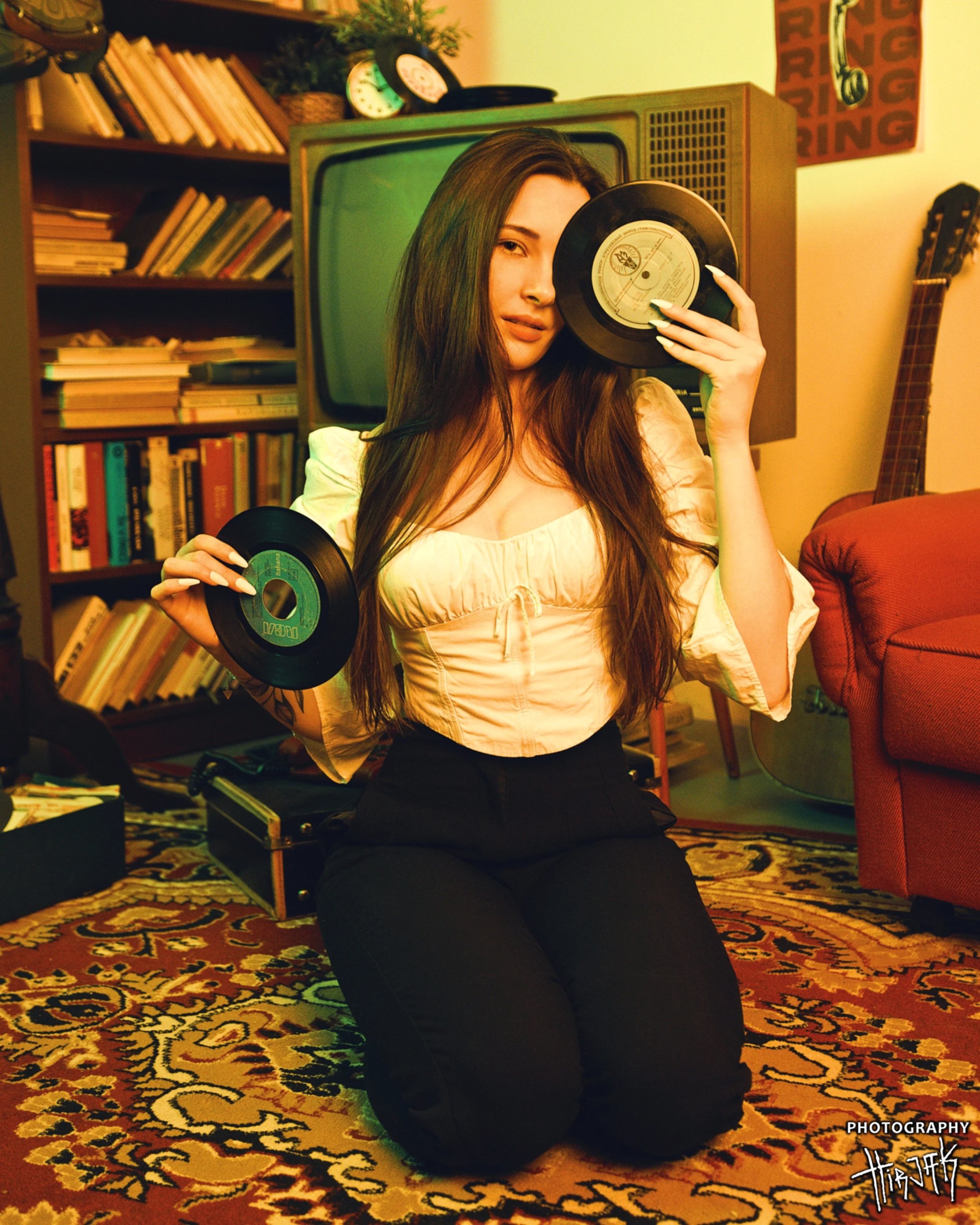 A young woman with long brown hair posing with vinyl records in a cozy, vintage-styled room. She is sitting on the floor, holding a record to her face and wearing a white puff-sleeve top and black pants. The background features a bookshelf filled wit