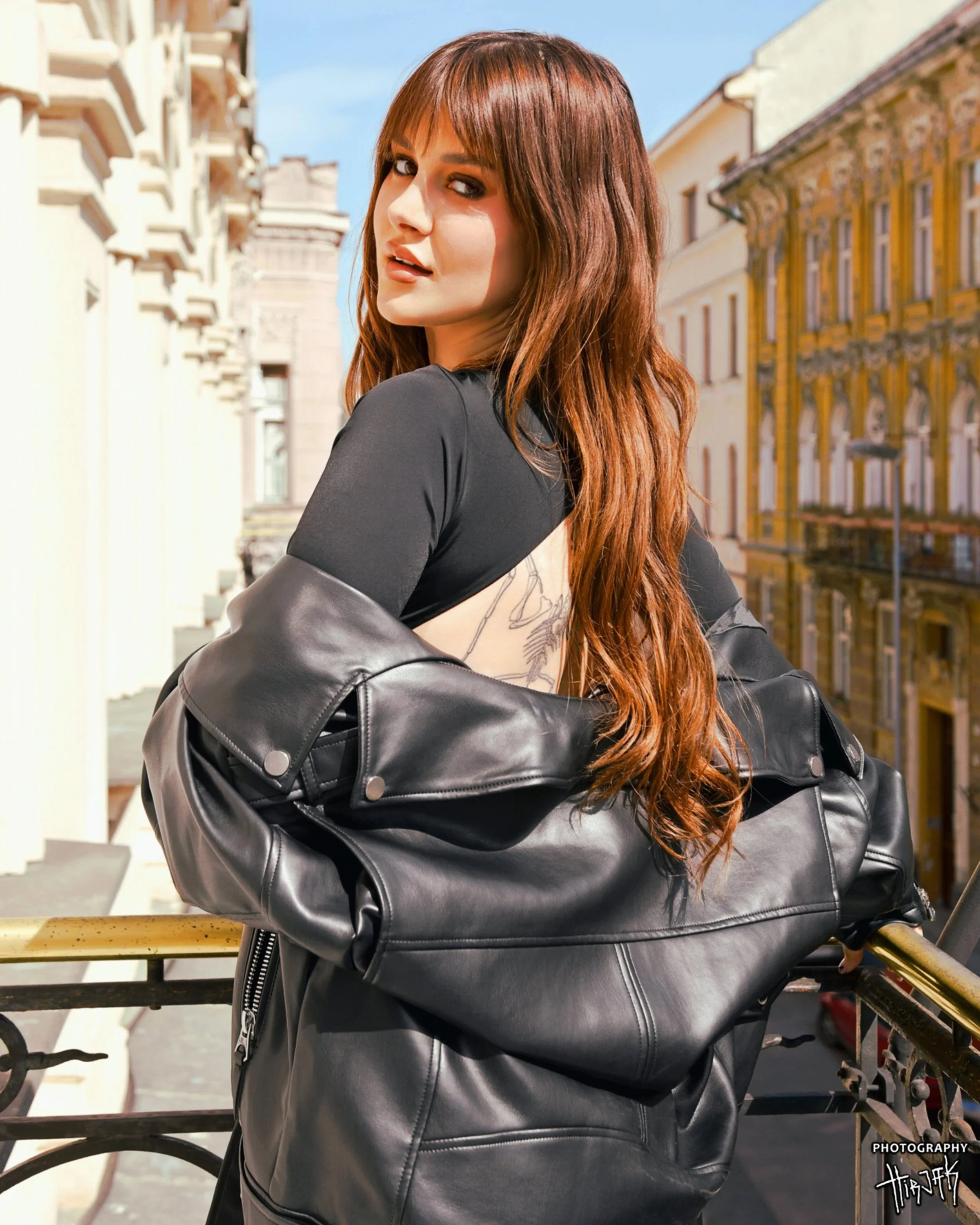 A young woman with long red hair and dark eye makeup posing outdoors near buildings with detailed architecture, wearing a black leather jacket, and leaning on a railing.