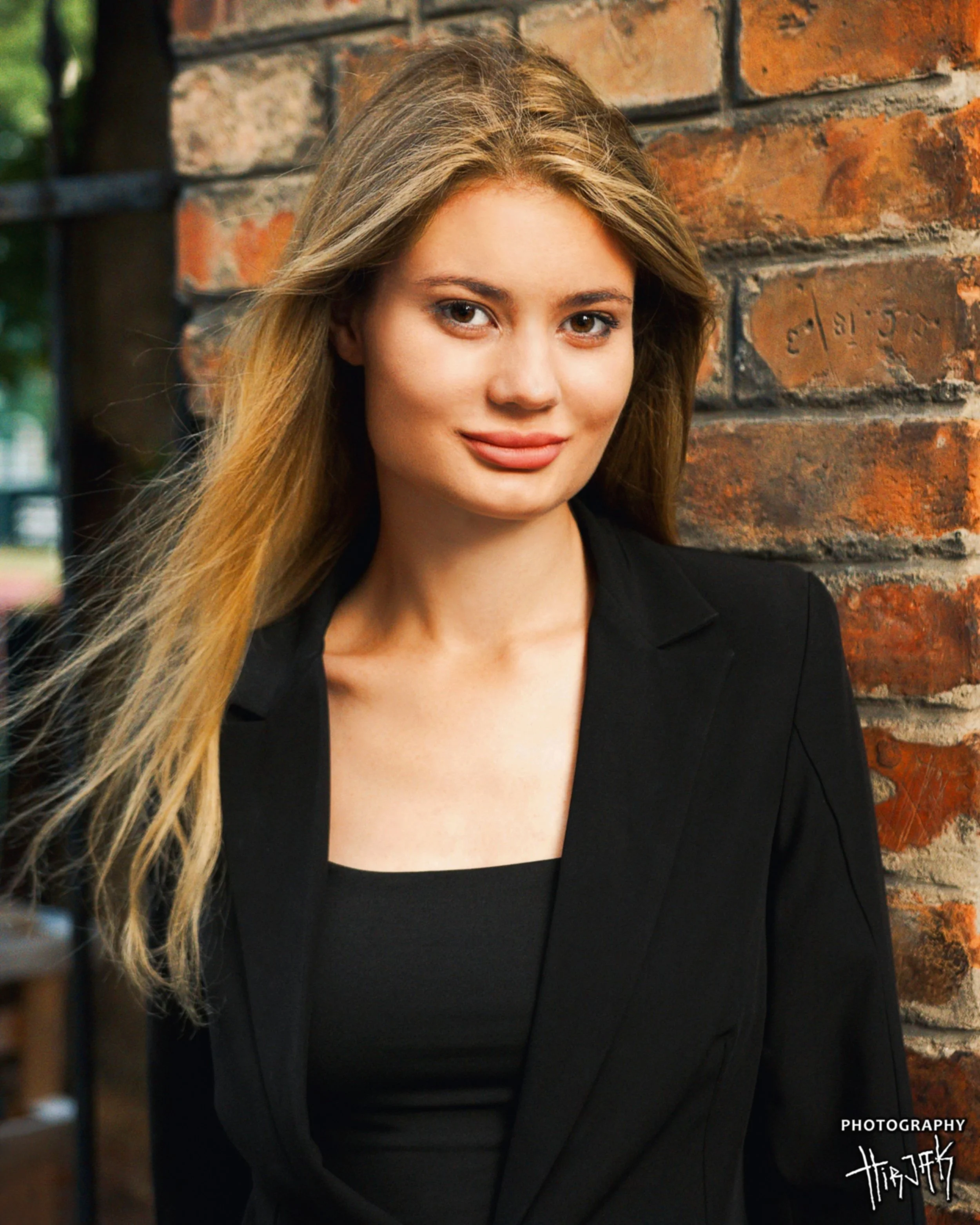 A young woman with long, light brown hair and brown eyes, wearing a black blazer and black top, standing against a brick wall. She has a slight smile, and the photo appears to be a professional portrait.
