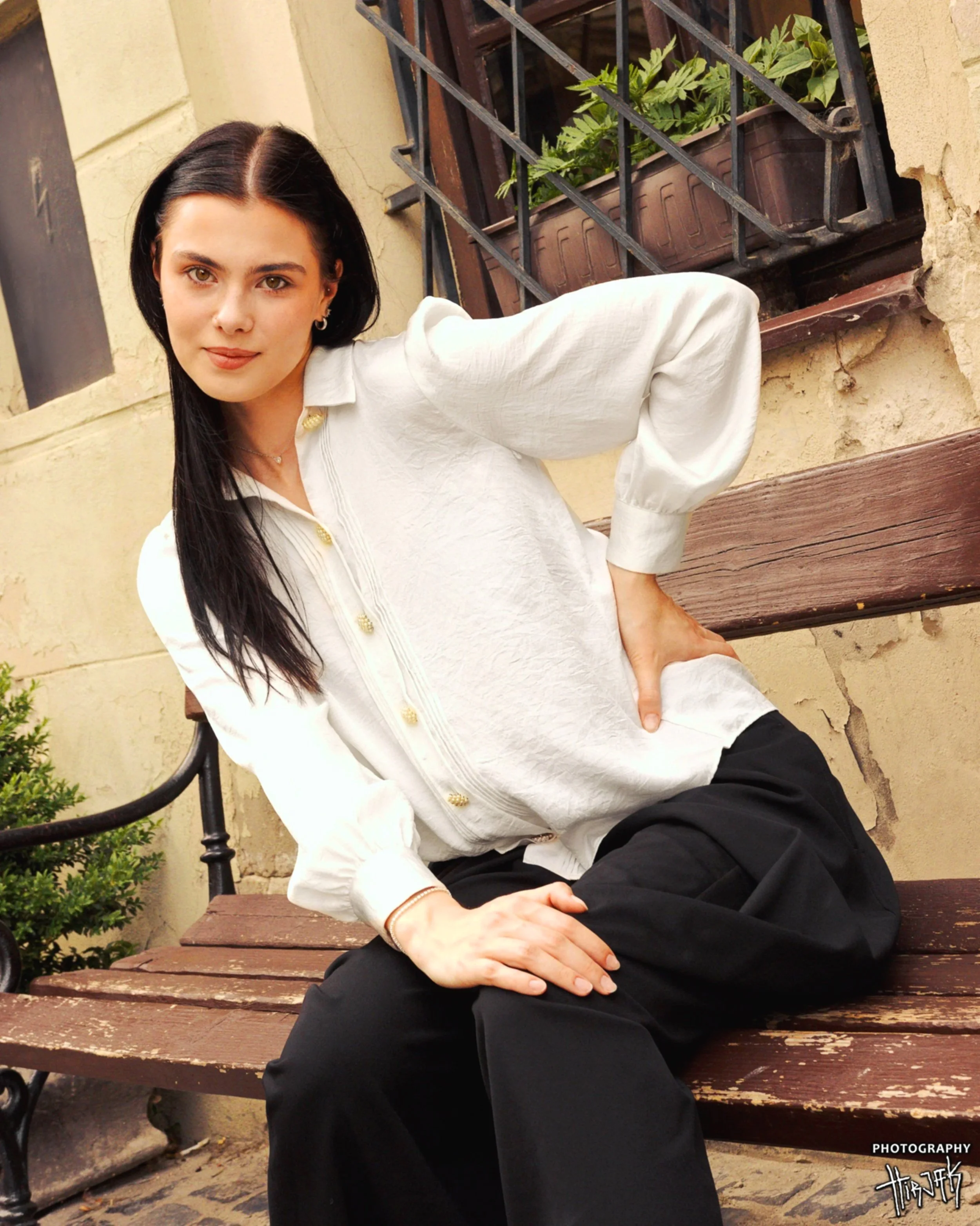 A woman with black hair and fair skin sitting on a wooden park bench, wearing a white blouse and black pants, with a background of a beige wall and window with plant pots.