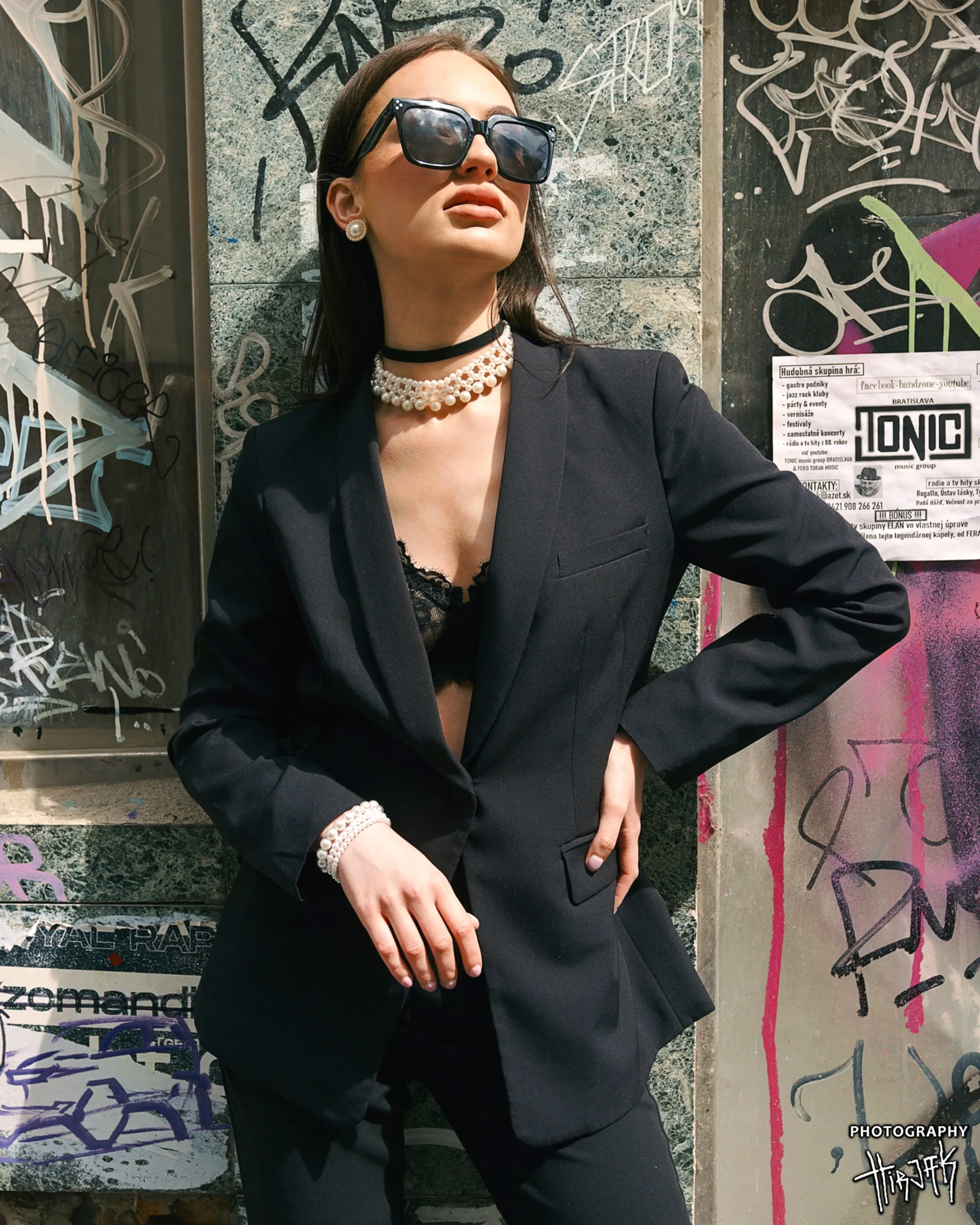 A woman with dark brown hair wearing large sunglasses, pearl earrings, a pearl necklace, a black blazer, and black pants, standing against a graffiti-covered wall on the street.
