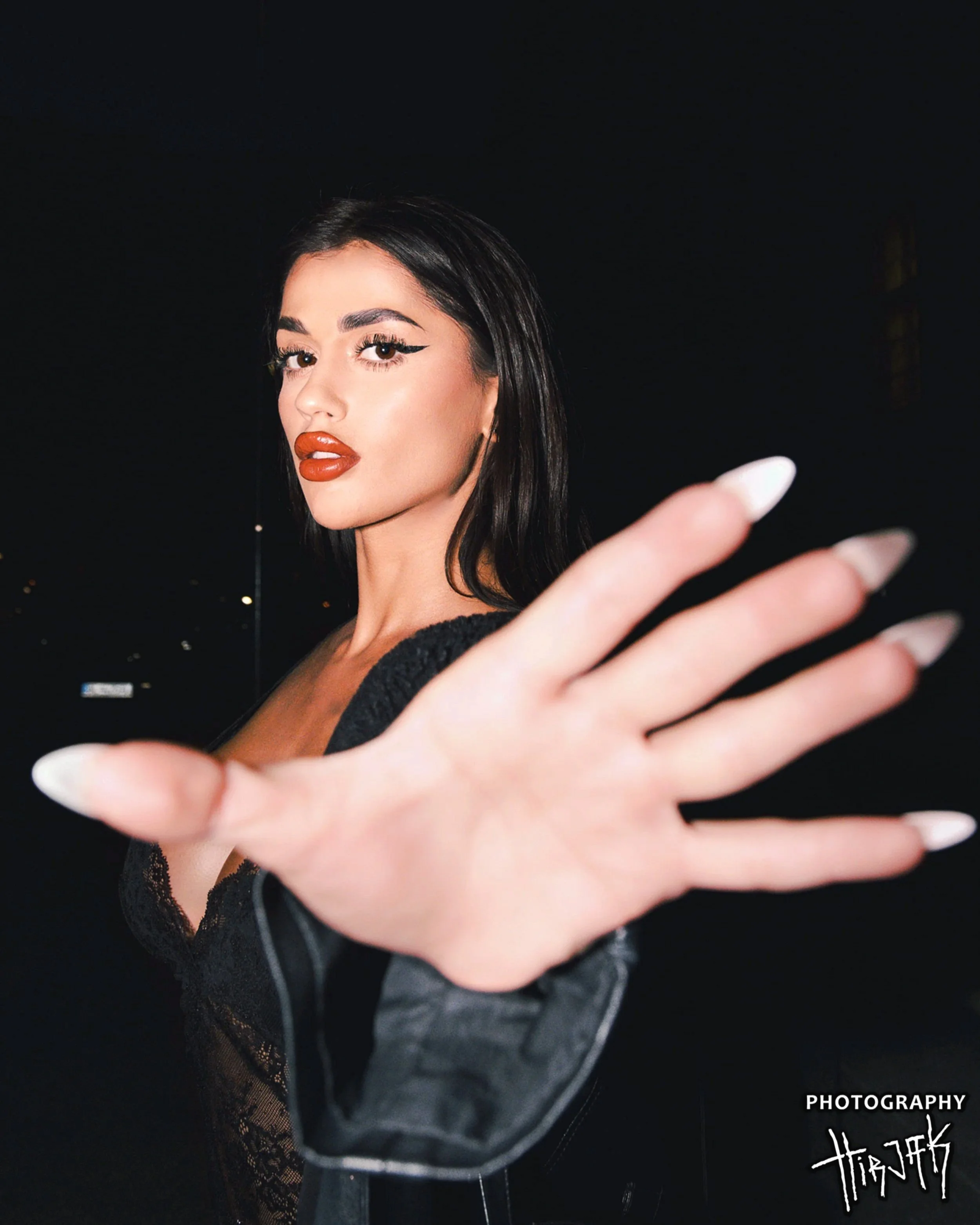 A woman with dark hair and makeup, wearing a lace top, extending her hand towards a camera in a night setting.