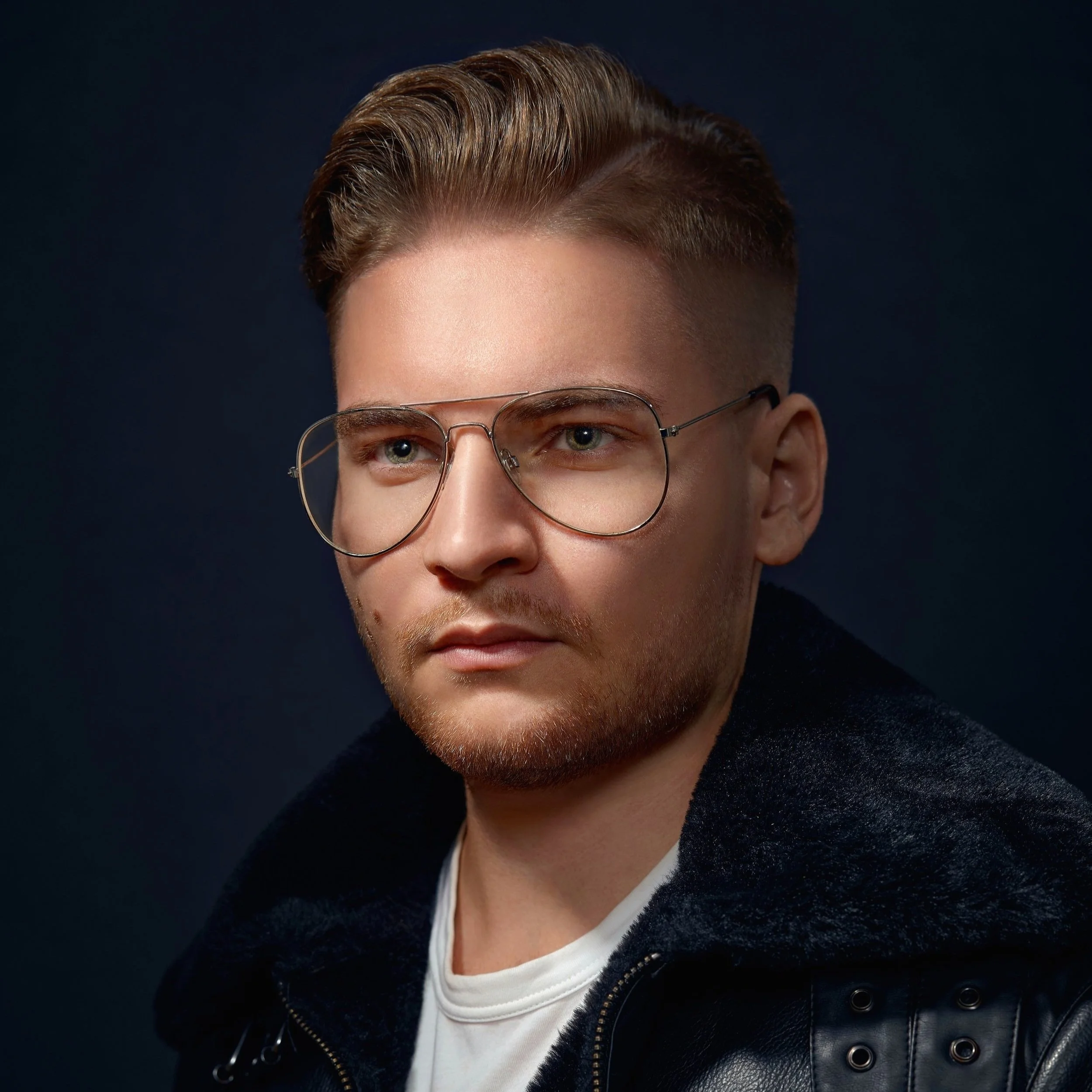 A young man with short, styled hair and wearing glasses, a white t-shirt, and a black jacket with a furry collar, against a dark background.