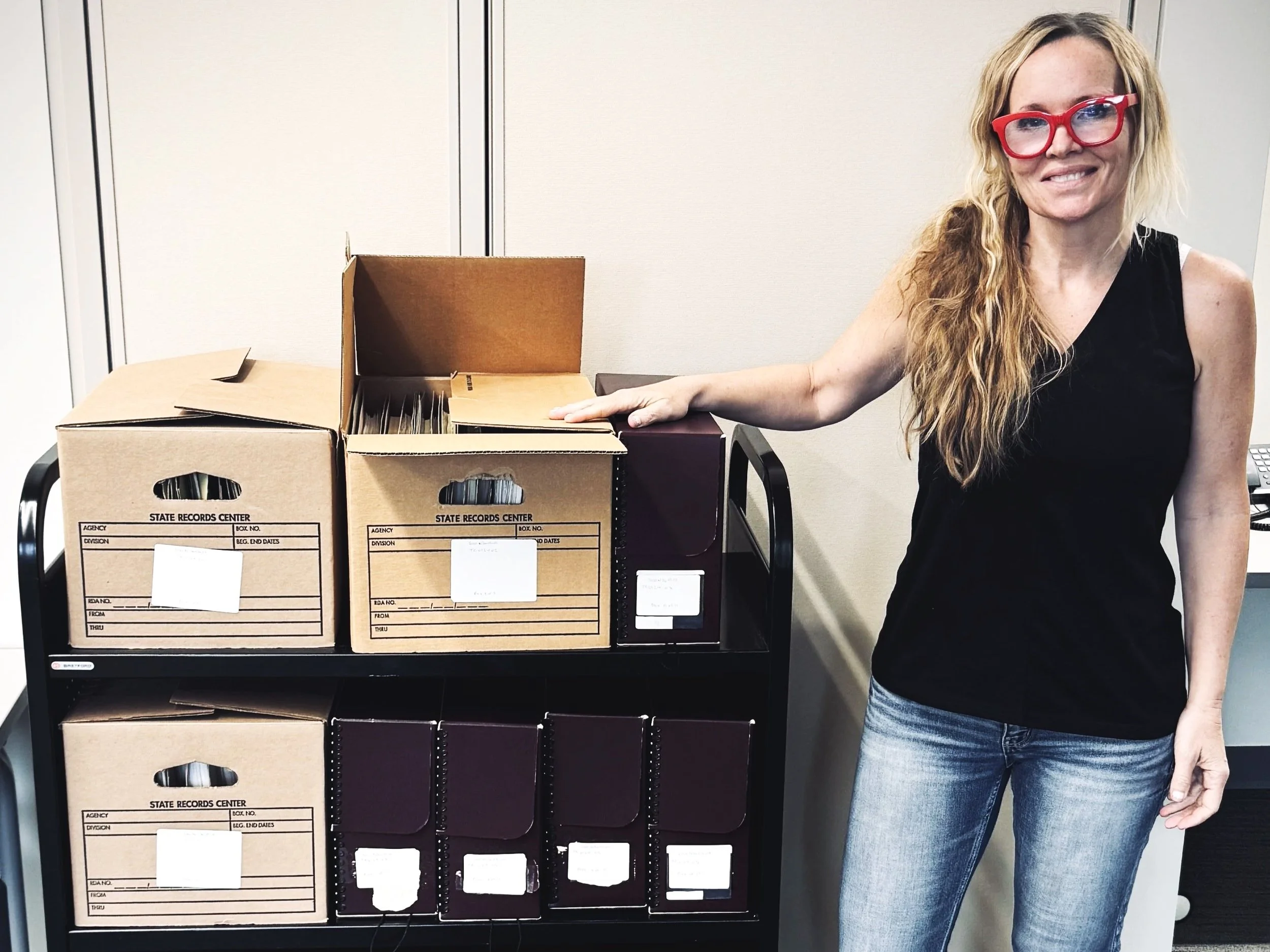 Historian and WWII researcher Erin Faith Allen stands beside a fully loaded archive cart holding three shelves of record boxes and binders at a state records center, mid-research.