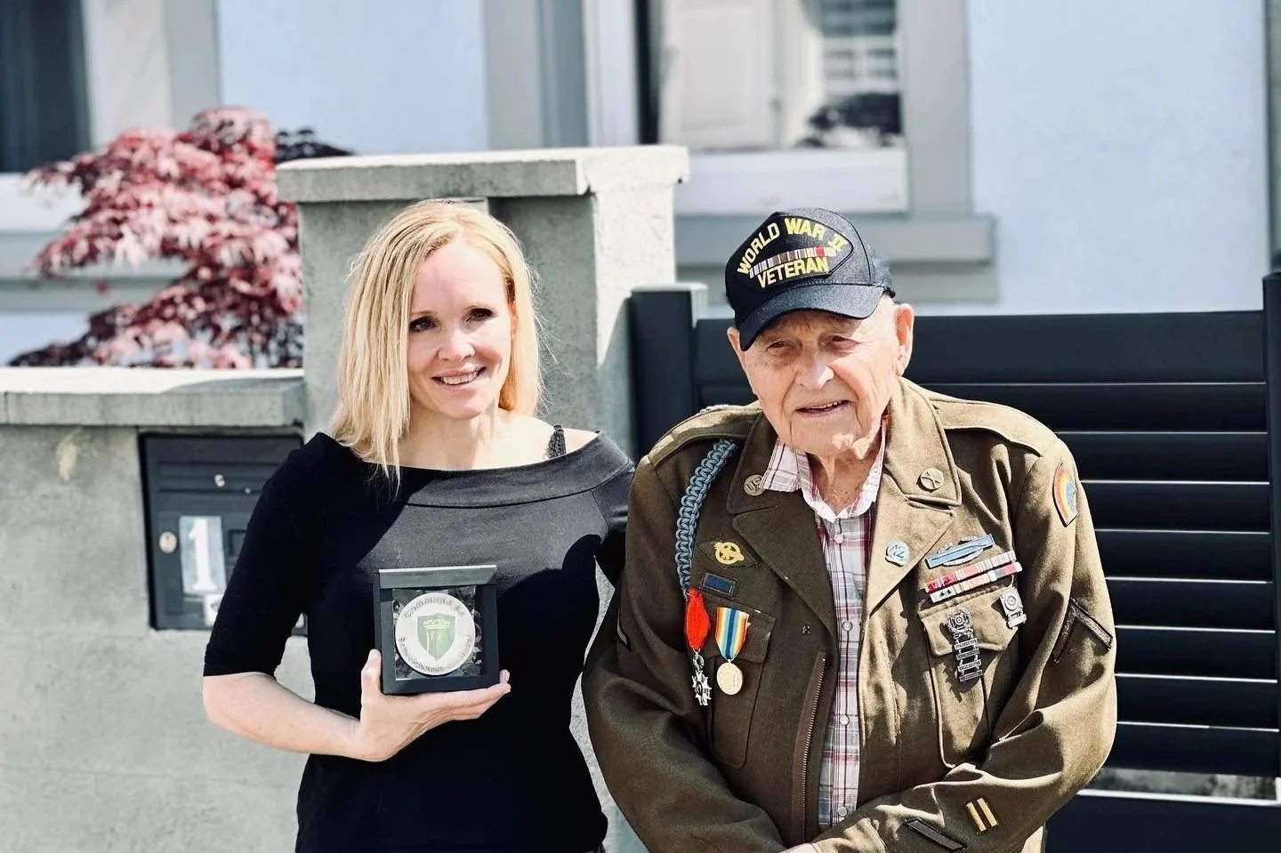 Historian Erin Faith Allen receiving honorary citizenship plaque from Schweighouse-sur-Moder, France, standing with 42nd Rainbow Division veteran Lockered "Bud" Gahs in front of the house where PFC Wayne Cruse was killed in January 1945.