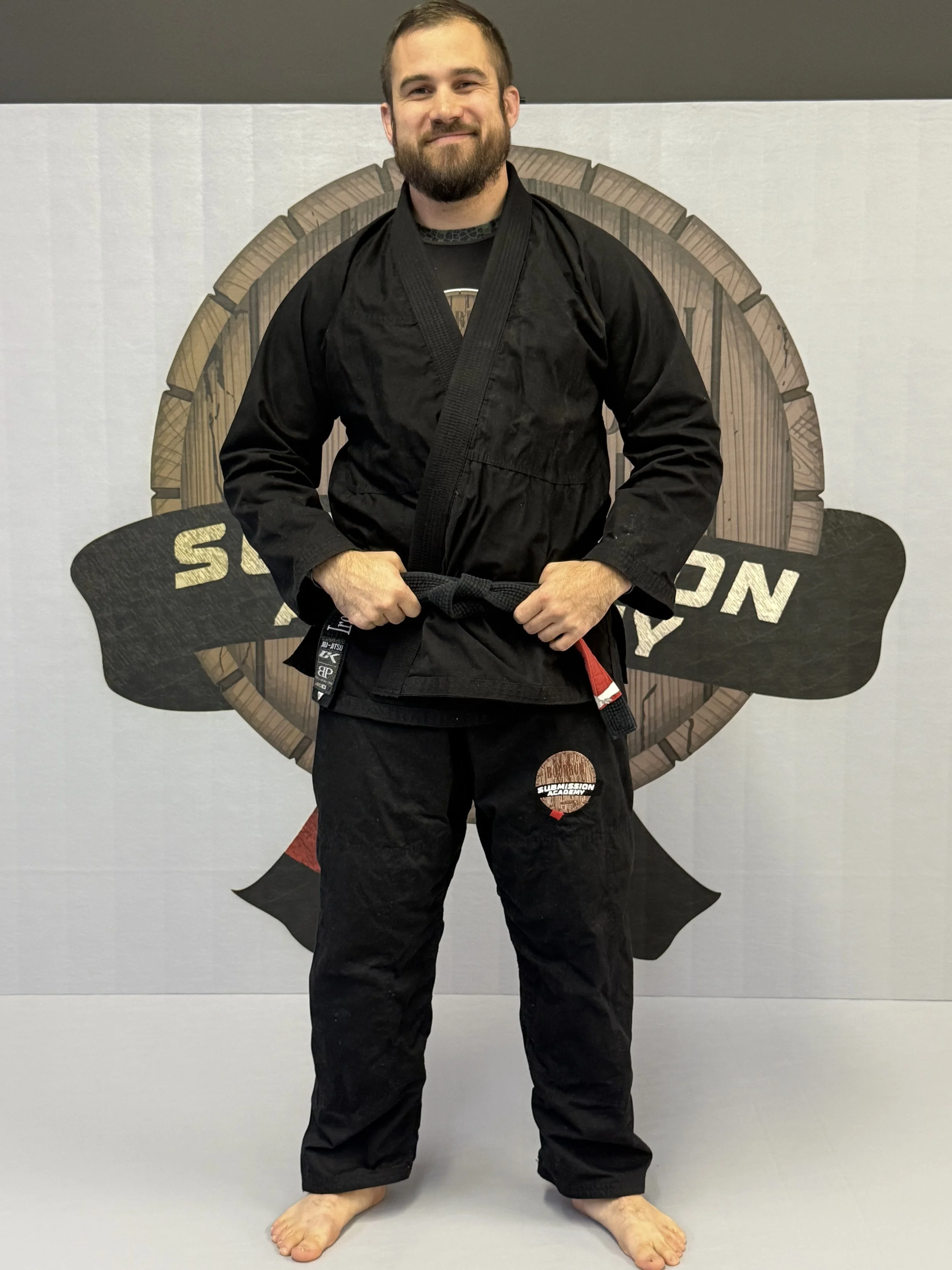 Two people practicing Brazilian Jiu-Jitsu on a blue mat, wearing black gis with belts. One person is attempting a sweep or guard technique on the ground, while the other is standing over them. Other practitioners are visible in the background.