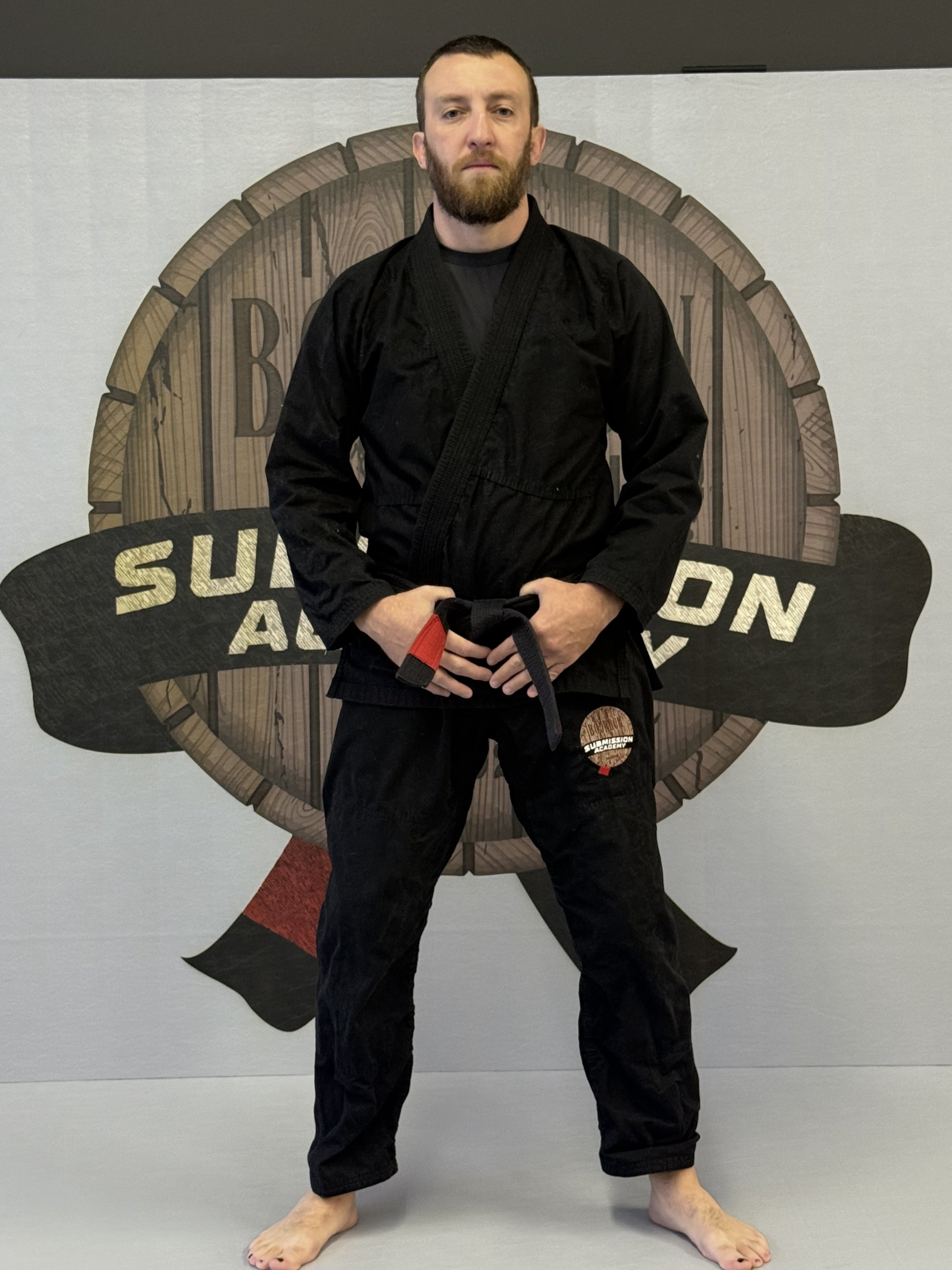 Two men wearing Brazilian Jiu-Jitsu gis and black belts standing on a mat in front of a plain wall, smiling and posing for a photo.