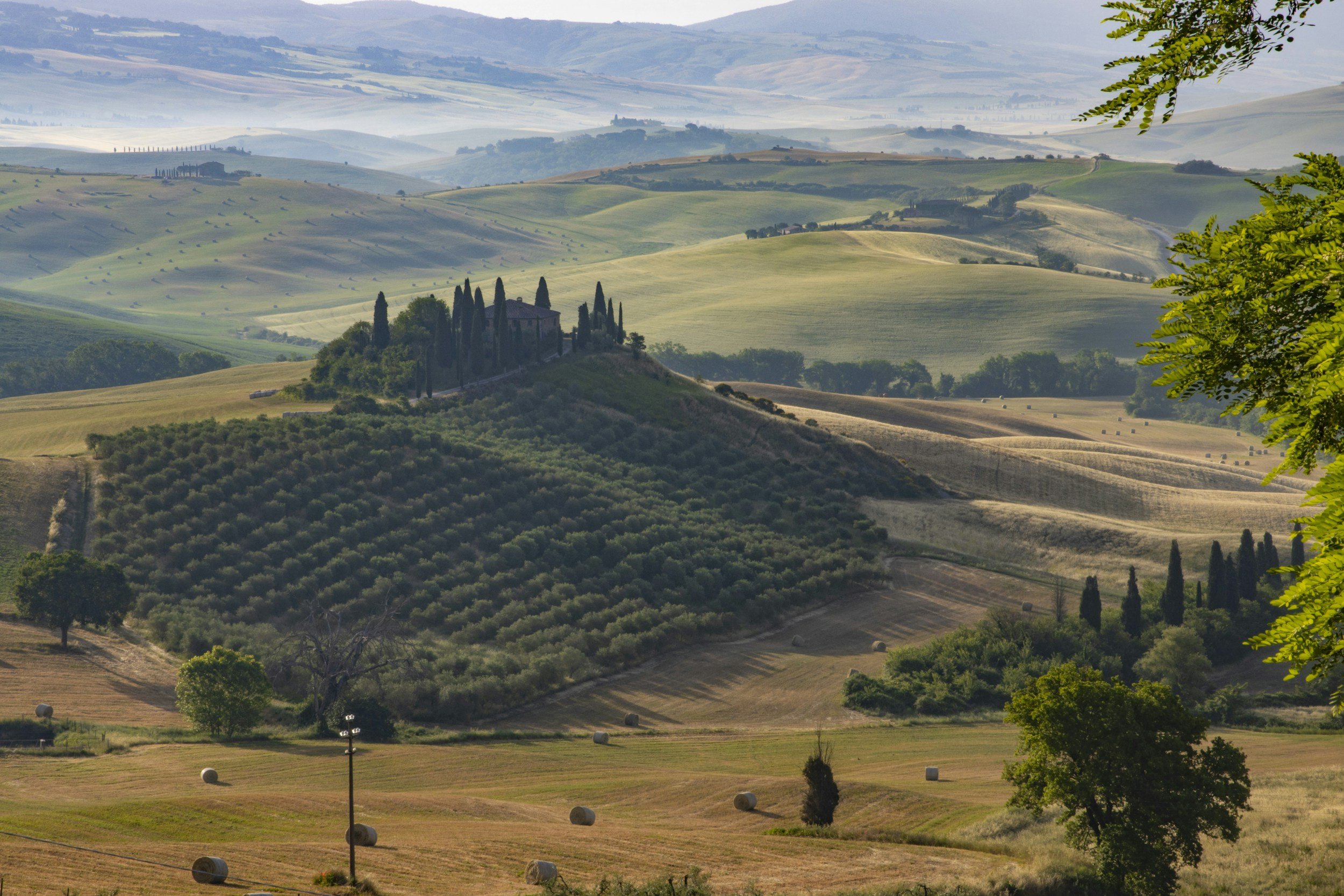 Scenic view of rolling hills and farmland with fields, trees, and hay bales, featuring a small house or villa surrounded by tall cypress trees on a hill in the Italian countryside.