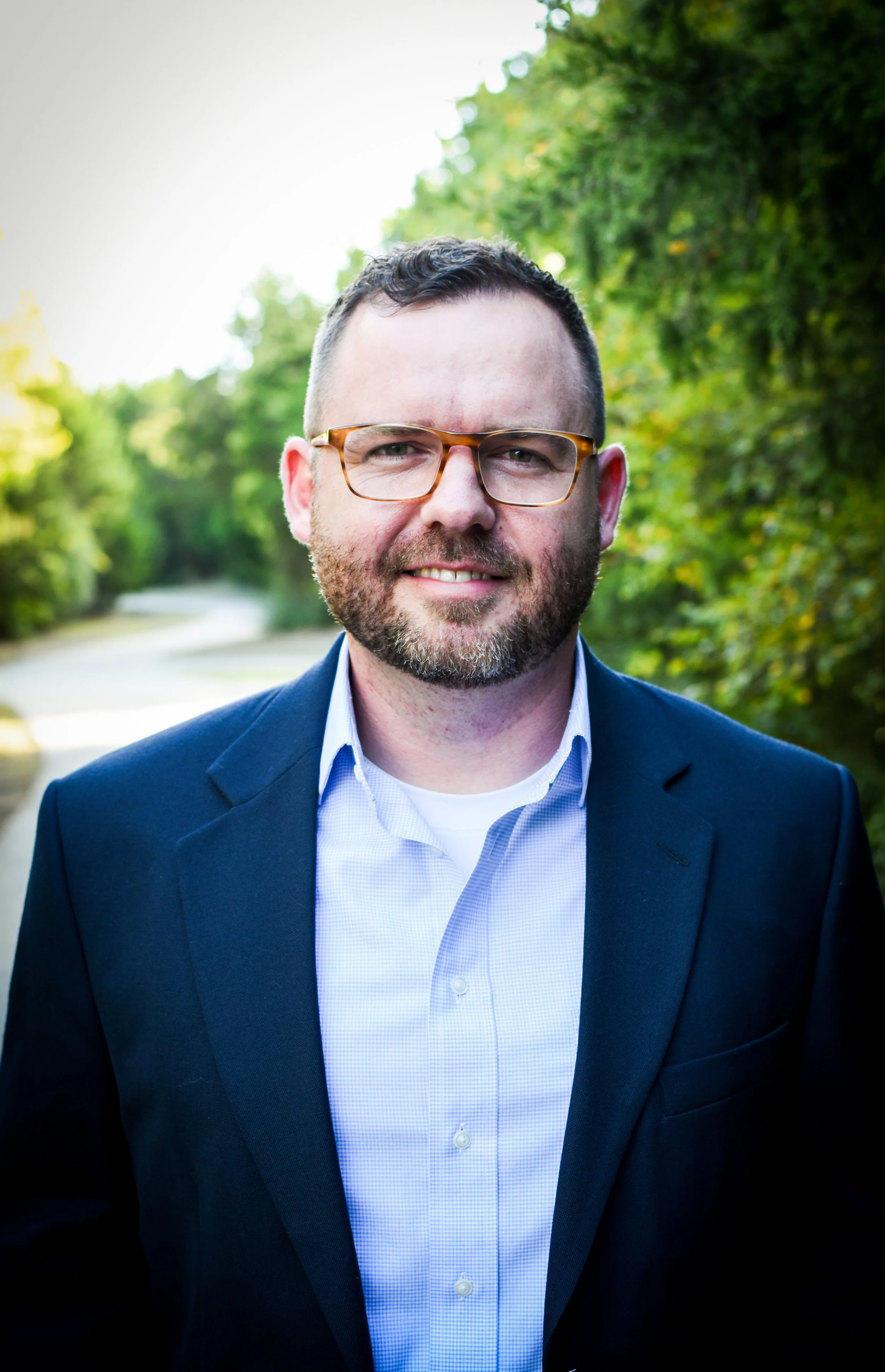 A man with glasses and a beard wearing a navy blue blazer and light blue shirt outdoors, smiling, with green trees in the background.