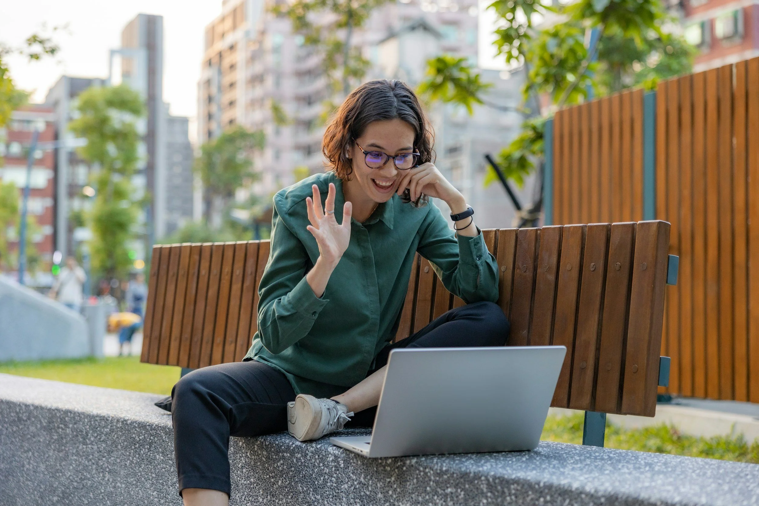 A woman with glasses, wearing a green shirt, sitting on a park bench outdoors, using a laptop, and talking on her phone, with city buildings and trees in the background.