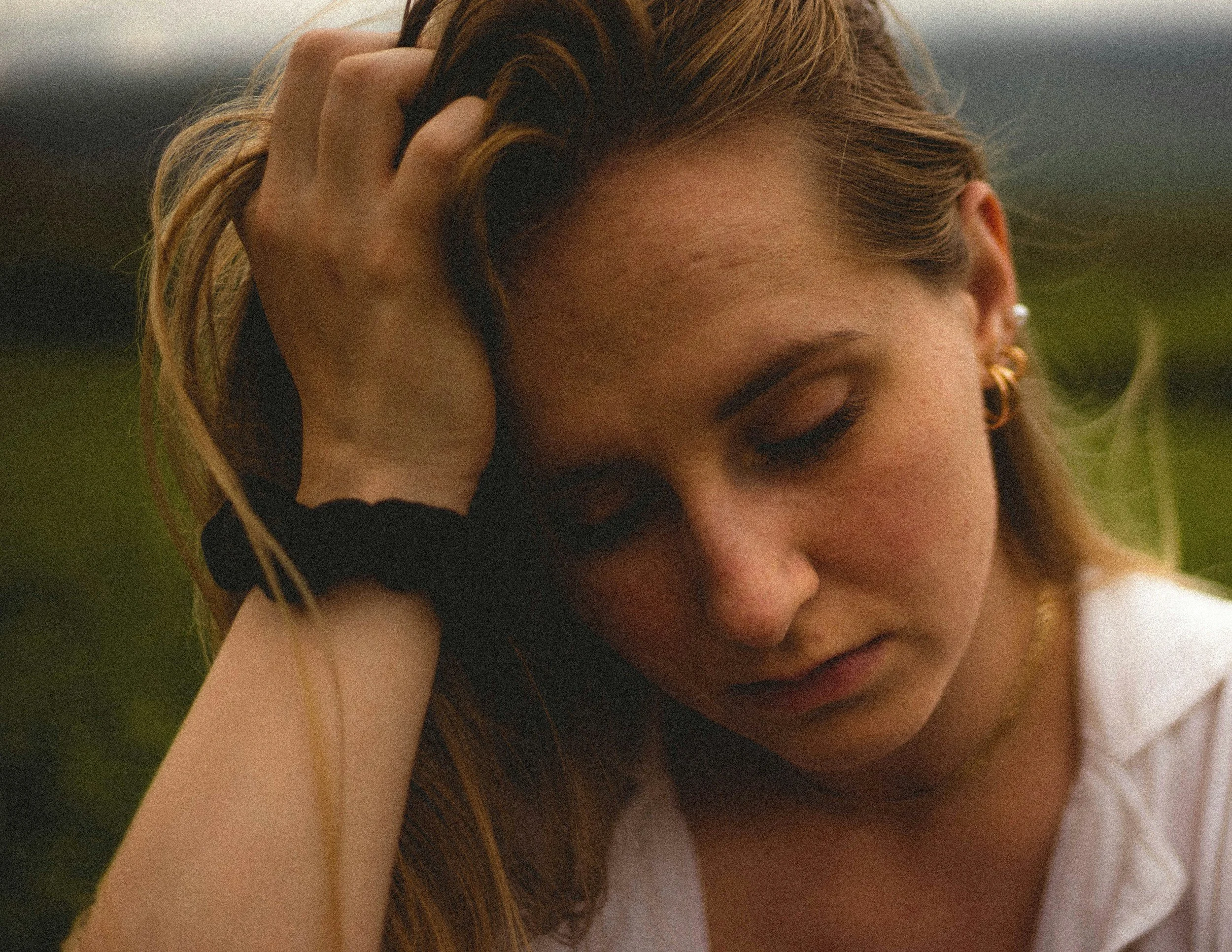 Close-up of a woman with light brown hair and earrings, resting her face with her eyes closed, hand on her head, outdoors with blurred green background.