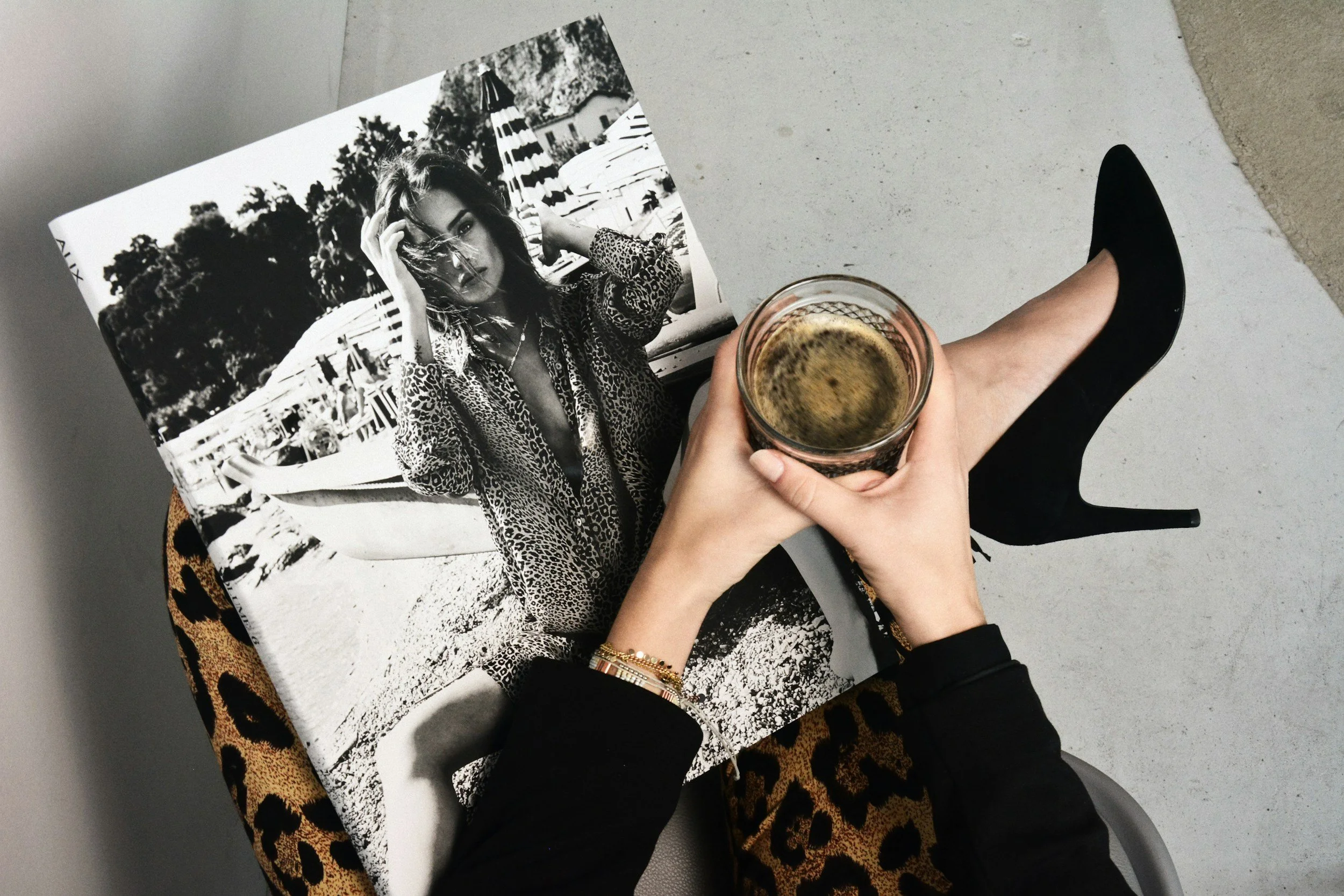 A person holding a glass of coffee or espresso, sitting at a leopard print chair with a black high heel boot, a magazine with a black-and-white photo of a woman in sunglasses and a leopard print coat, on a concrete floor.