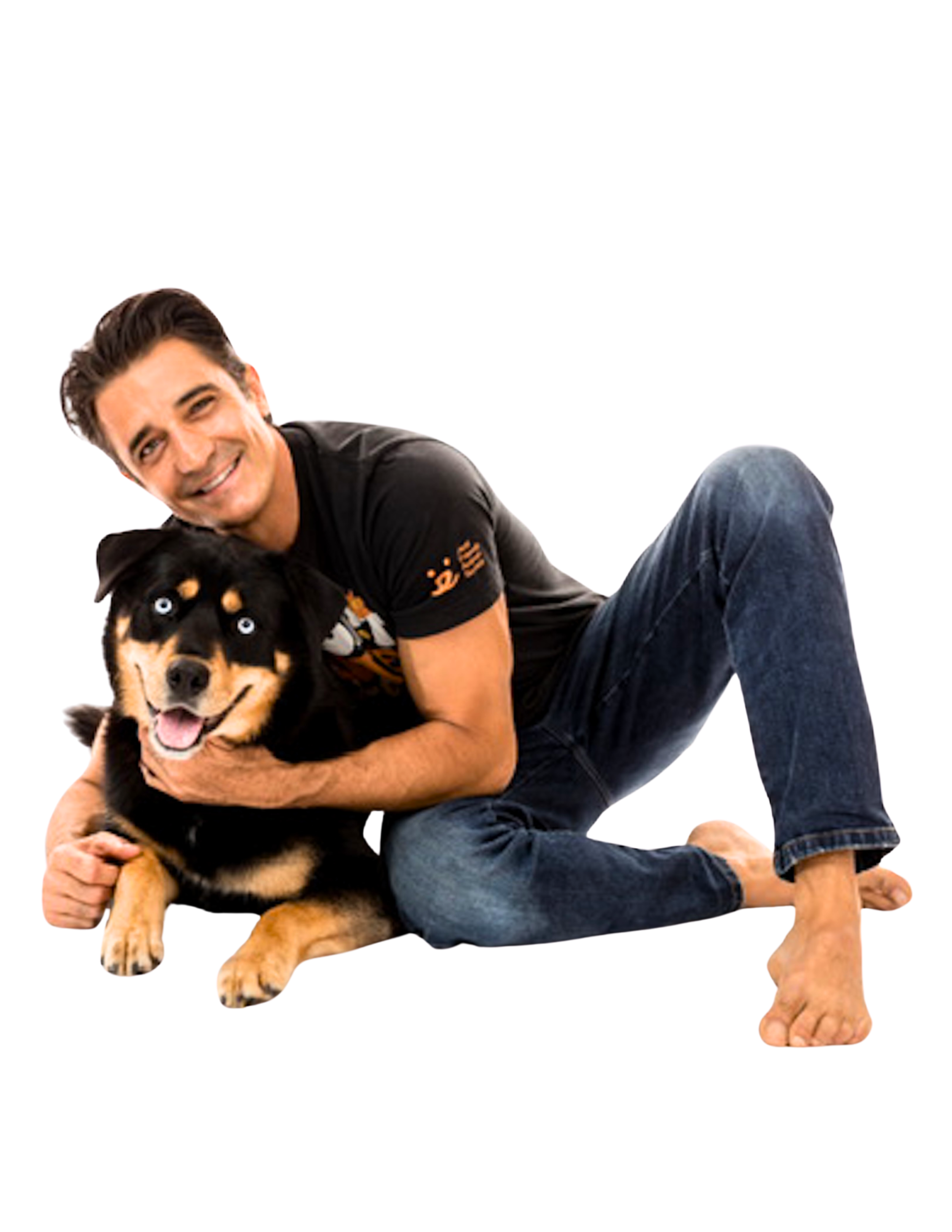 A man with dark hair, wearing a black t-shirt and jeans, smiling and hugging a happy black and tan dog with cloudy blue eyes, sitting on a plain background.