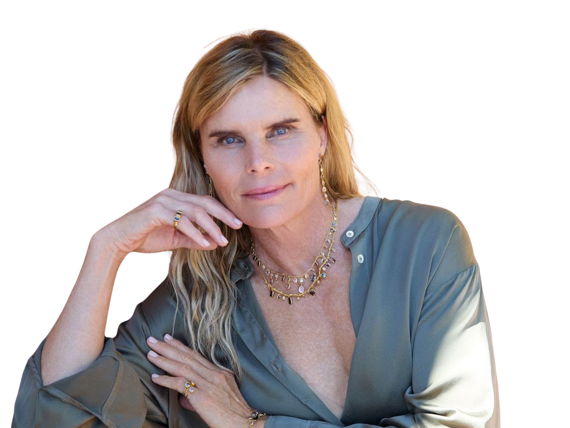Portrait of a middle-aged woman with blonde hair, blue eyes, wearing a shiny grey blouse and layered jewelry, posing with her hand resting on her face and her other arm crossed.