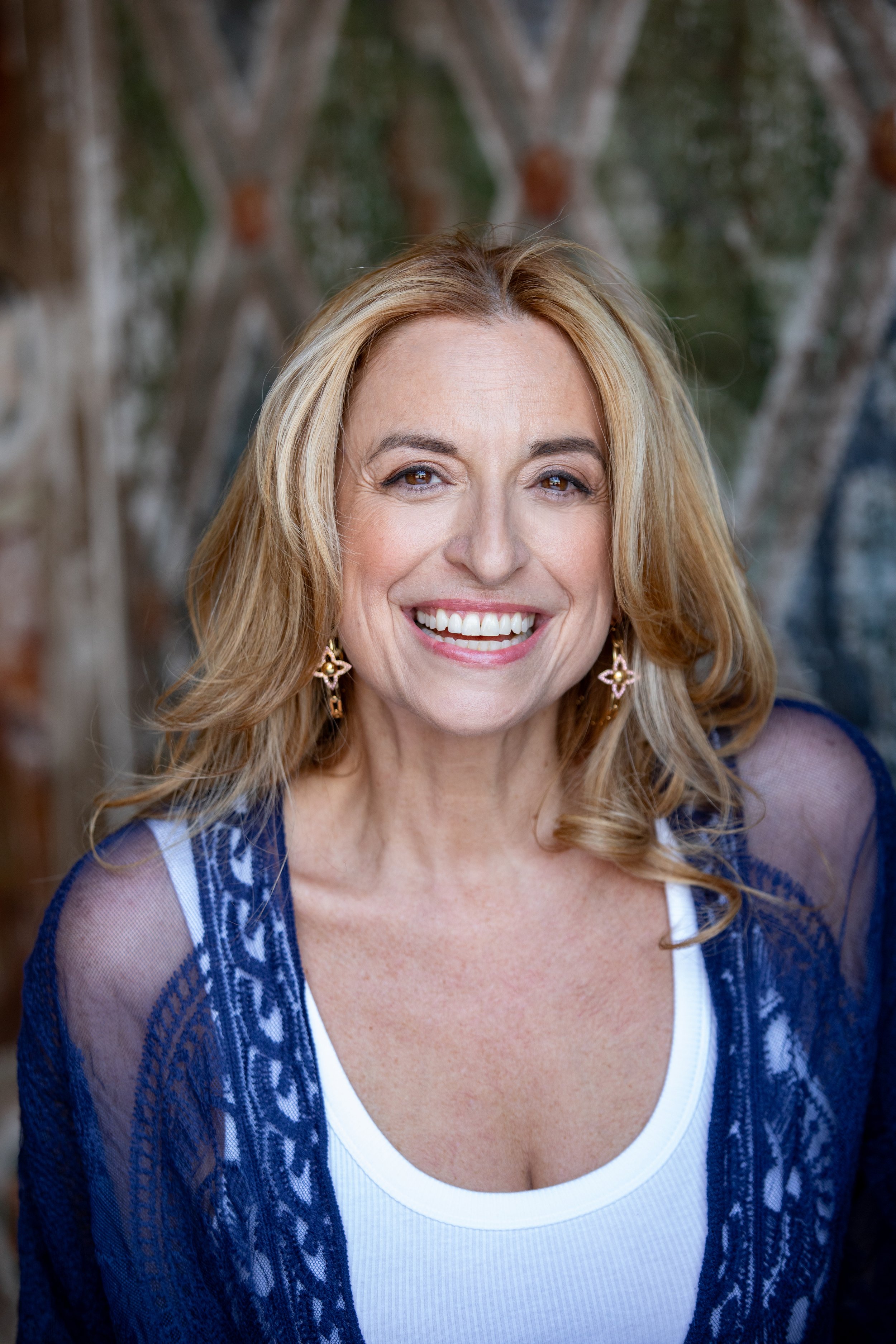 A smiling woman with blonde hair, wearing earrings and a white top with a blue lace cover-up, outdoors with trees in the background.