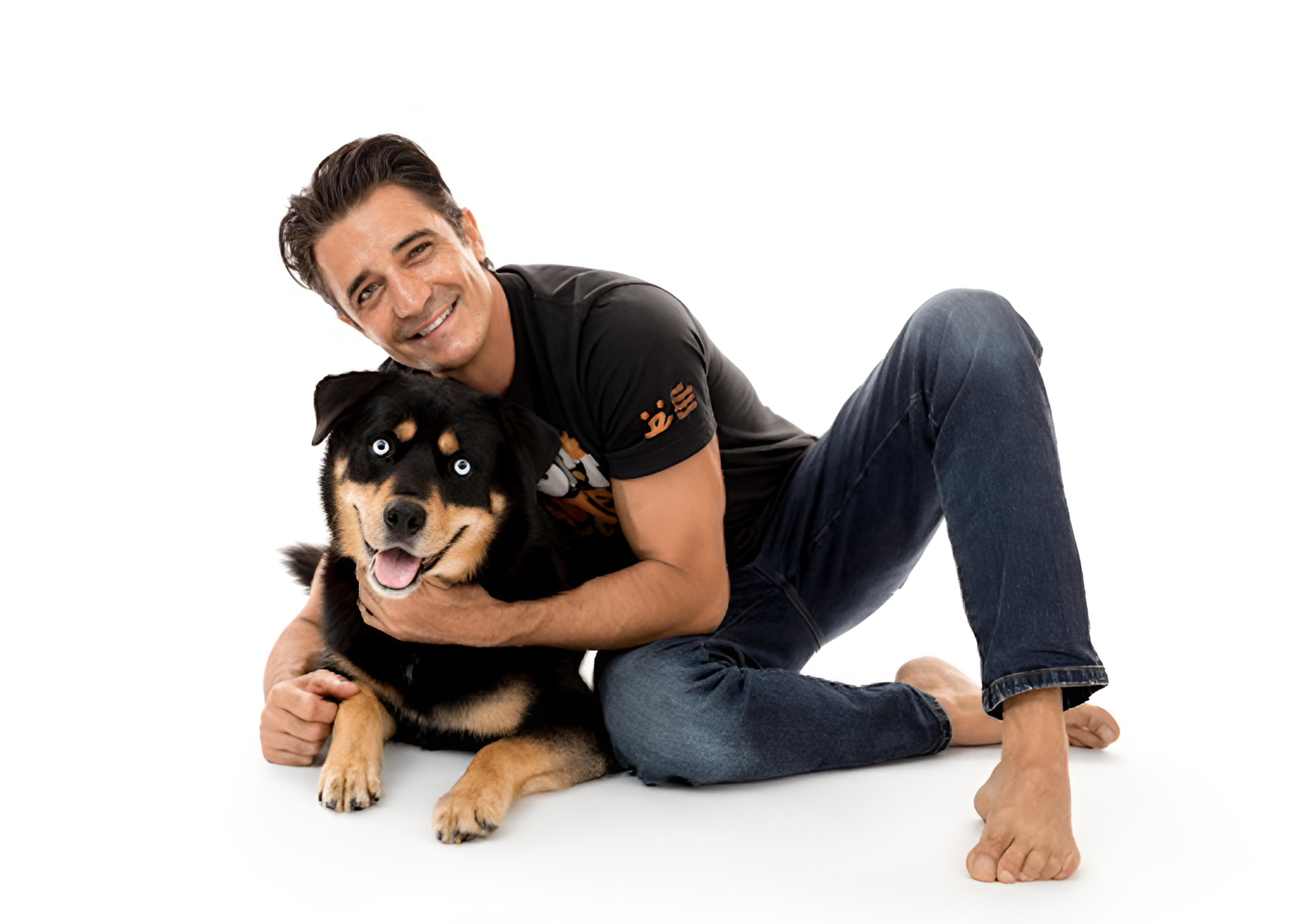 A man with dark hair smiling and sitting on the floor, hugging a happy black and tan dog with blue eyes, in front of a plain white background.