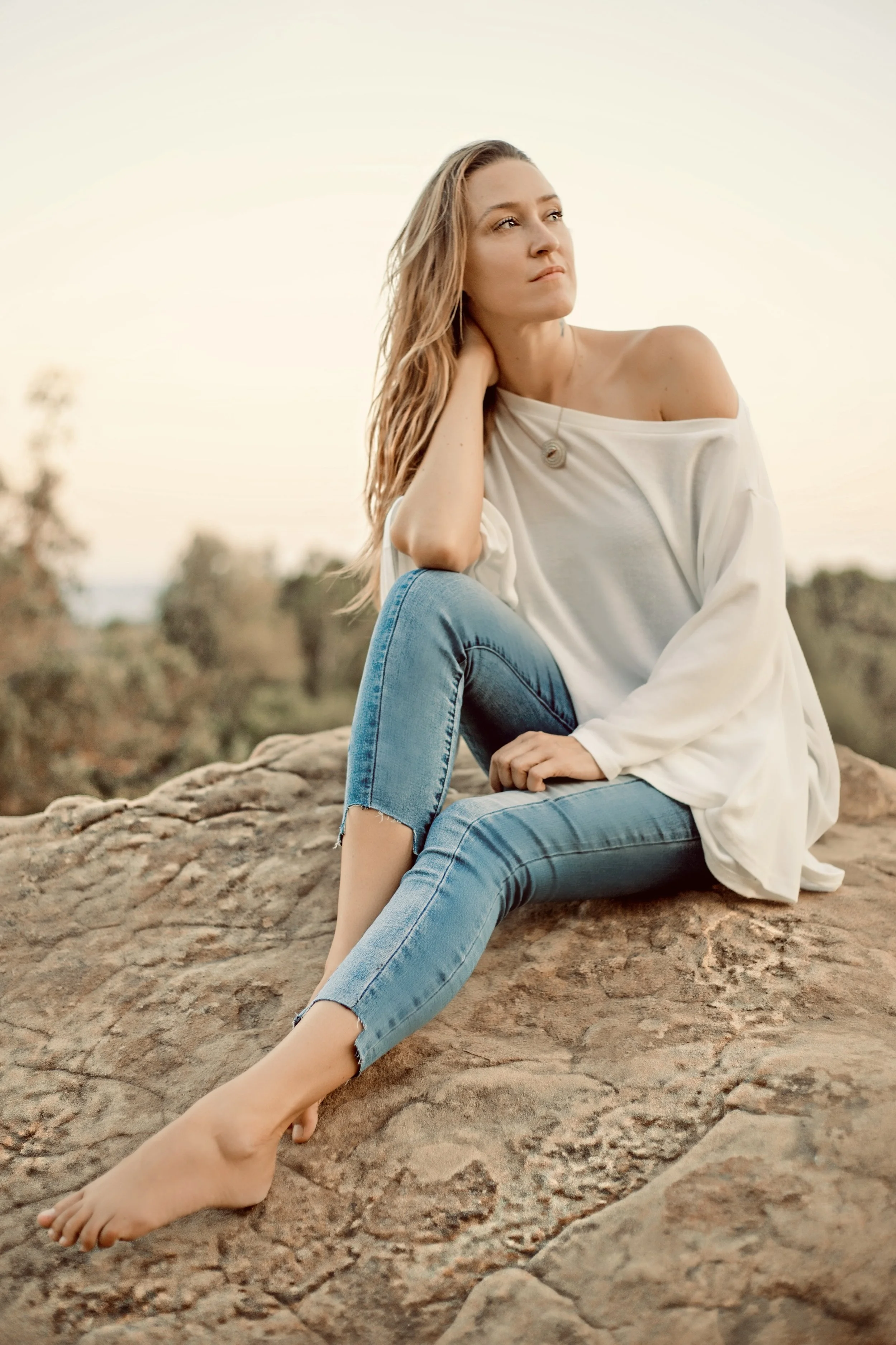 A woman with long blonde hair sitting on a large rock, wearing a loose off-shoulder white top and ripped blue jeans, looking contemplative in an outdoor setting at sunset.