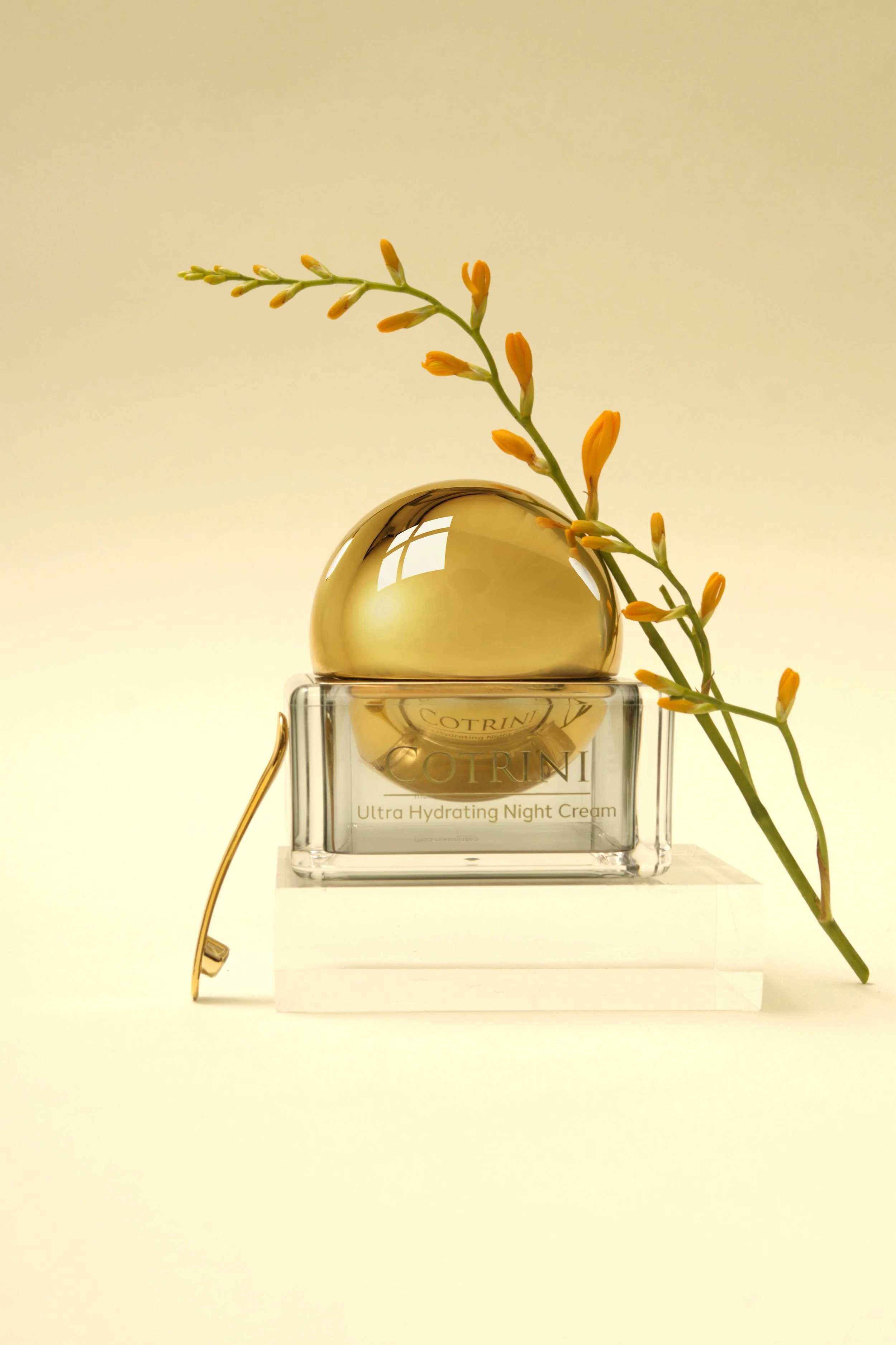 A jar of Cotrin Ultra Hydrating Night Cream with a gold spoon or applicator and a sprig of yellow-orange flowers, all on a clear acrylic block with a plain, light background.