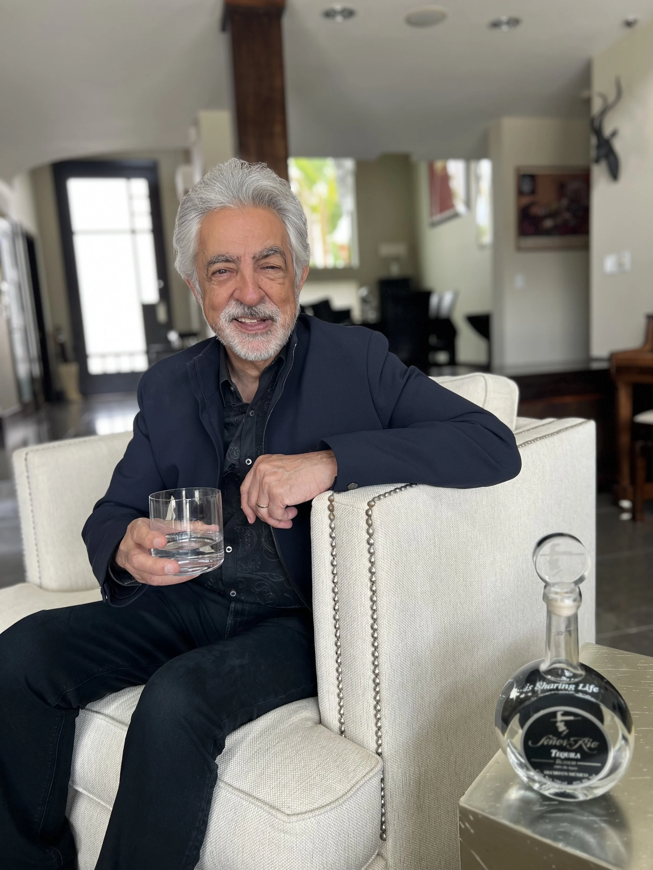 An older man with gray hair and a beard sitting on a cream-colored upholstered armchair, smiling, wearing a dark jacket, holding a glass of water, with a bottle of tequila on a silver table beside him in a bright, modern living room.