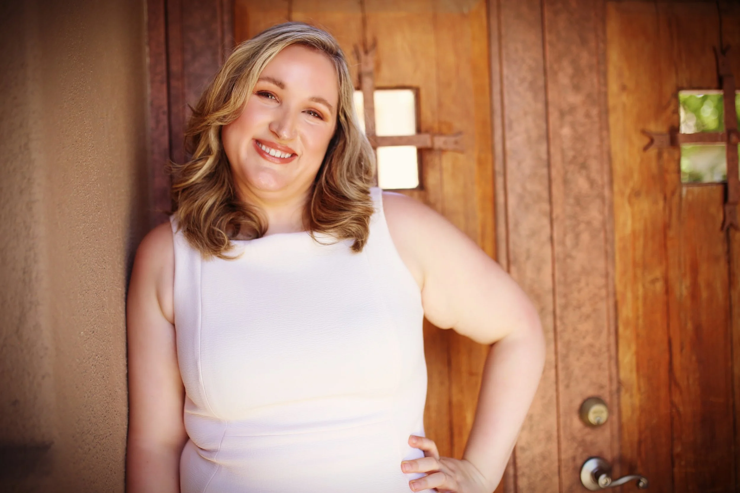 A woman with blonde hair smiling while leaning against a wooden wall with small windows, wearing a white sleeveless top.