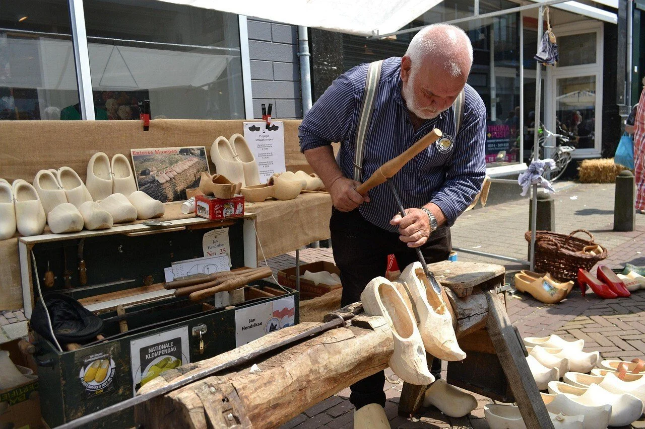 Un homme âgé sculpte des sabots en bois dans un marché en plein air. Un sabotier. Autour, des sabots en bois terminées et en cours de fabrication sont exposées.