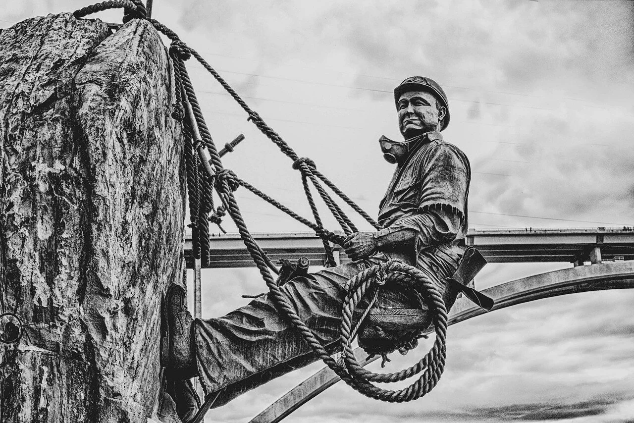 Statue en bronze d'un mineur avec casque, attaché à une roche artificielle, avec un pont en arrière-plan, en noir et blanc.