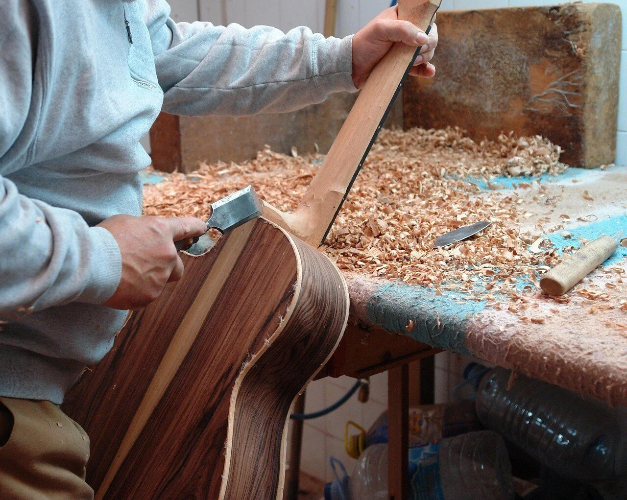 Un artisan en train de sculpter une guitare en bois dans un atelier, entouré de copeaux de bois.