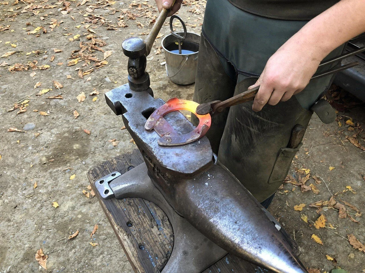 Un forgeron travaille sur un fer à cheval chauffé à la forge, dans un atelier en plein air, avec des feuilles mortes au sol.