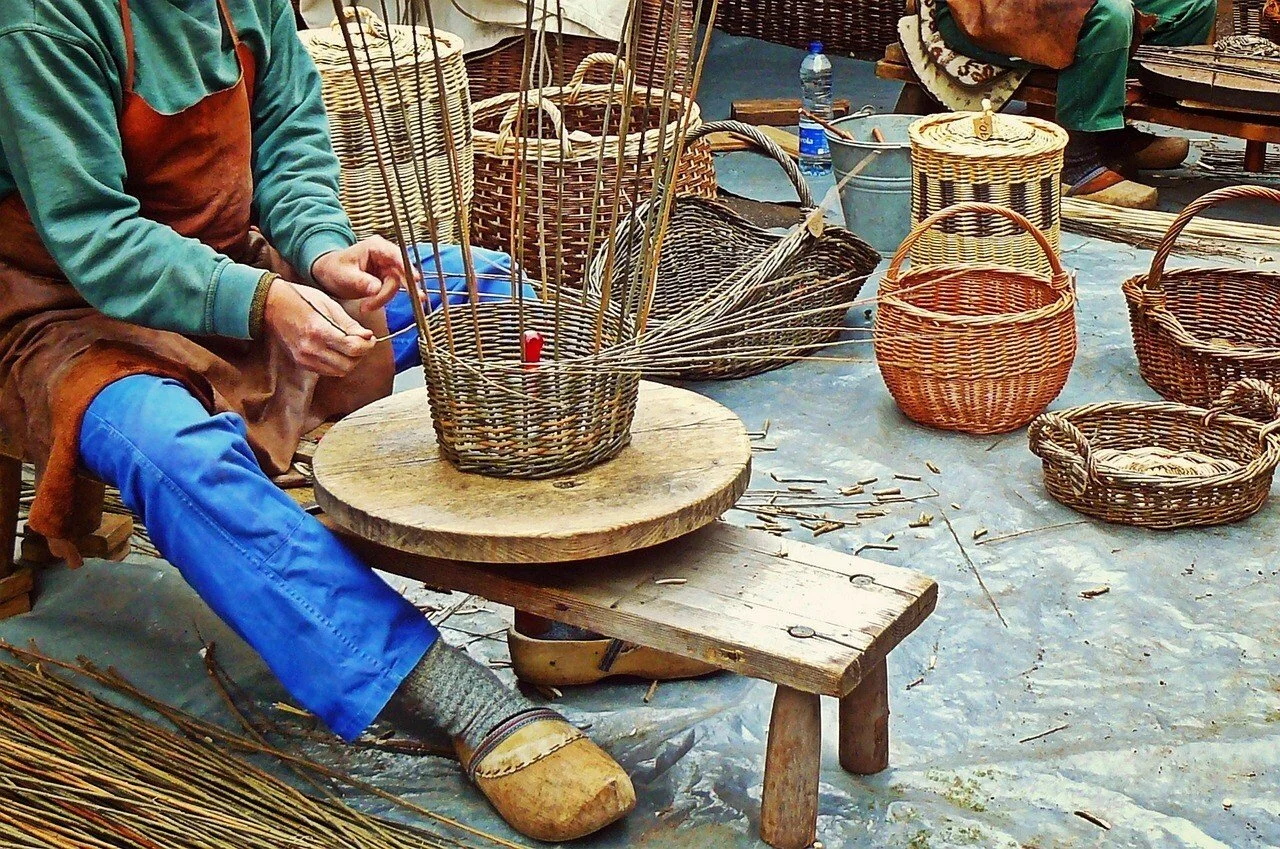 Une personne tisse un panier en osier à la main, autour d'une table en bois, avec plusieurs paniers de différentes tailles et formes sur le sol. Tisserant.