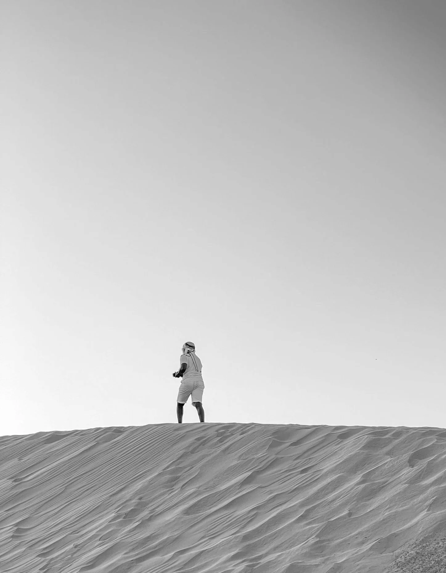 A person wearing a hat and shorts standing on a sand dune in a desert landscape with a clear sky.
