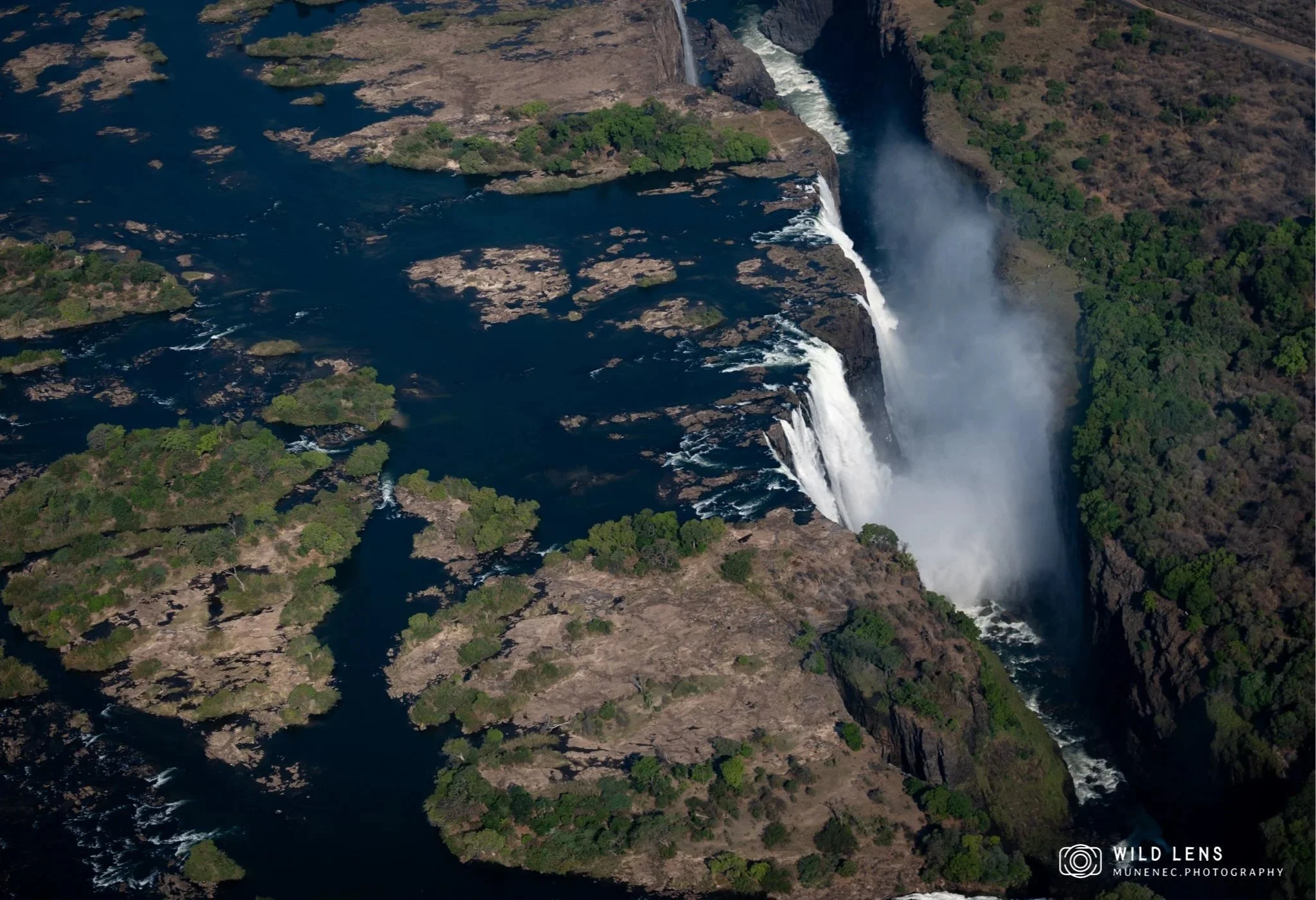 Aerial view of a waterfall cascading into a deep canyon with surrounding green vegetation and rocky terrain.