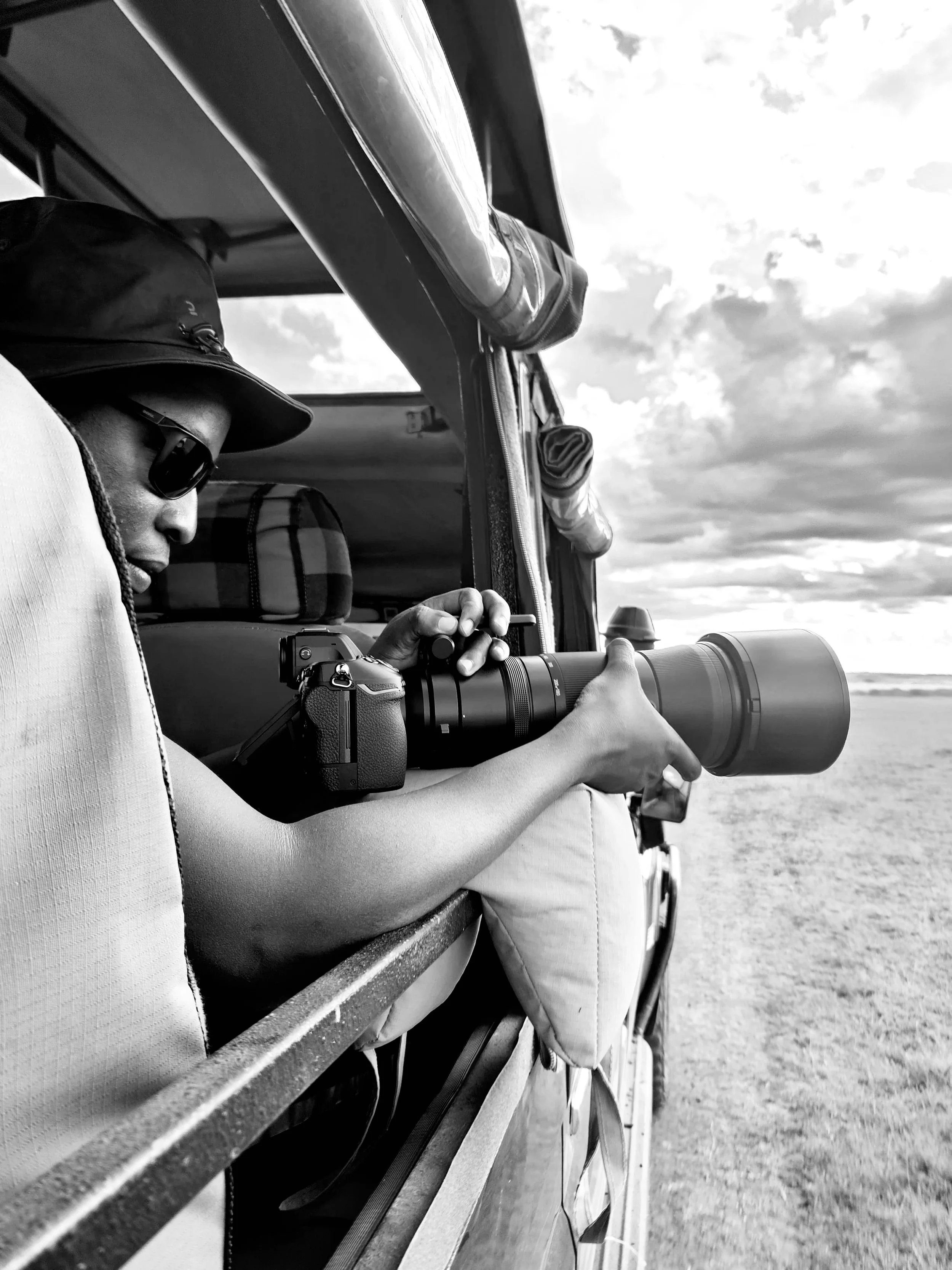 A person wearing a hat and sunglasses is holding a camera with a large telephoto lens out of the window of a vehicle, aiming at the landscape outside.