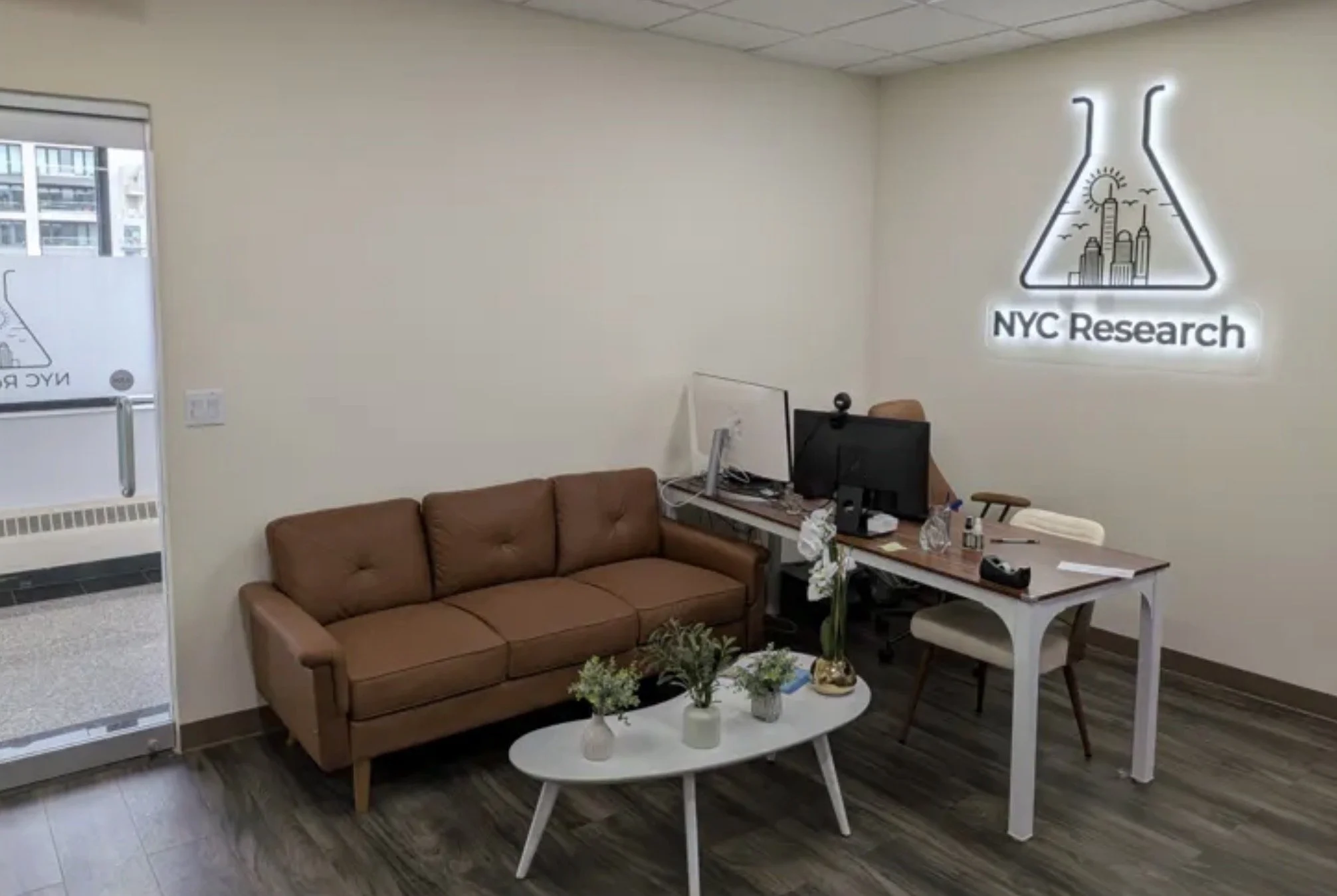 An office with a brown leather sofa, a white coffee table with white vases and flowers, a desk with two computer screens, and a wall sign reading 'NYC Research' with a cityscape and lab flask logo.