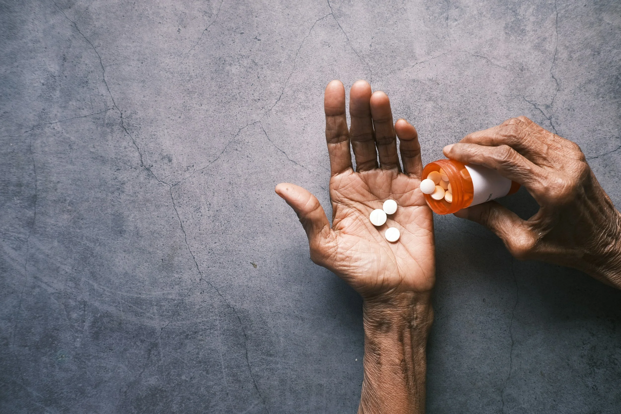 Close-up of an elderly person's hand dispensing pills from an orange bottle onto the palm of their other hand on a gray cracked surface.