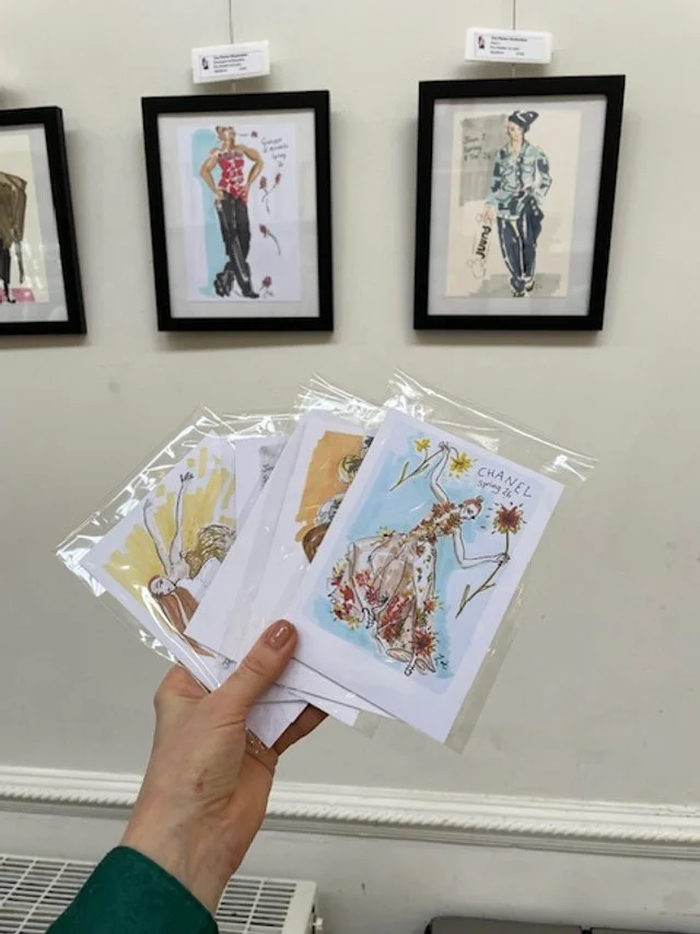 Zoe Parker Illustration's hand holding a variety of postcards for sale and framed fashion illustration prints.