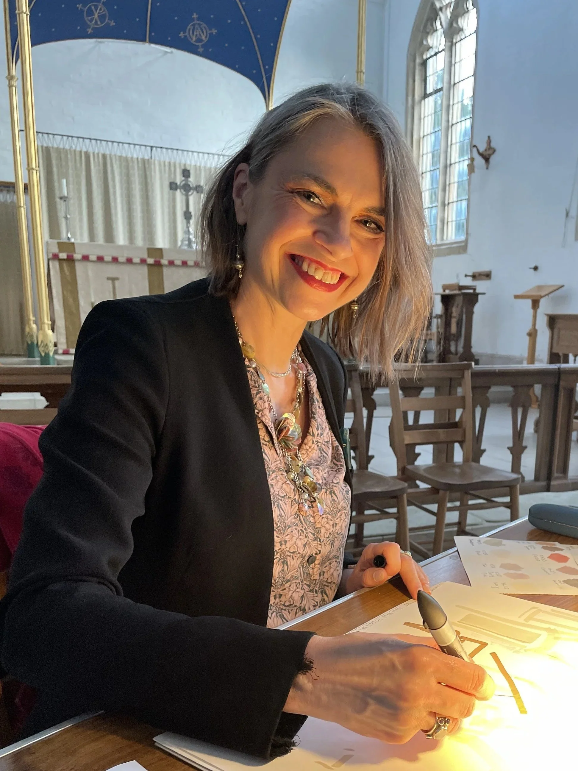Zoe Parker Illustration is creating a live wedding illustration in a church. She is smiling at her desk at the back of the church. She is creating live guest illustrations  and live portraits of the guests and the newly-weds. She is using promarkers.