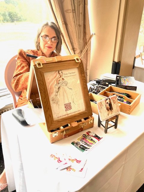 Zoe Parker illustration creating a live wedding illustration of guests at an Indian wedding. She is seated at a desk creating a drawing using ProMarker pens. She has a desk easel on the table in front of her, she is wearing an orange suit and concent