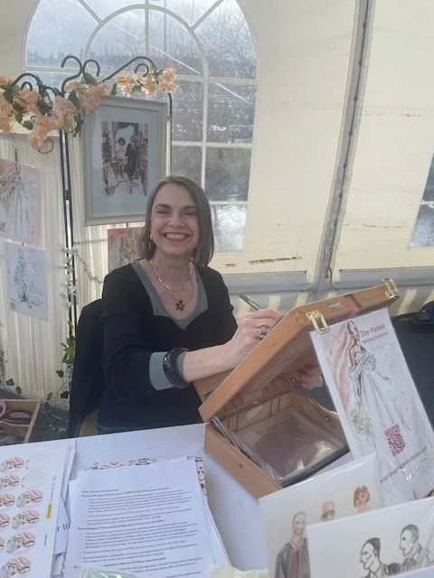 Zoe Parker Illustration seated at her table during Bluewater Signature Wedding Show arranged by County Wedding Events. She is smiling at the camera and drawing on her desk easel. Behind her are examples of her wedding illustrations