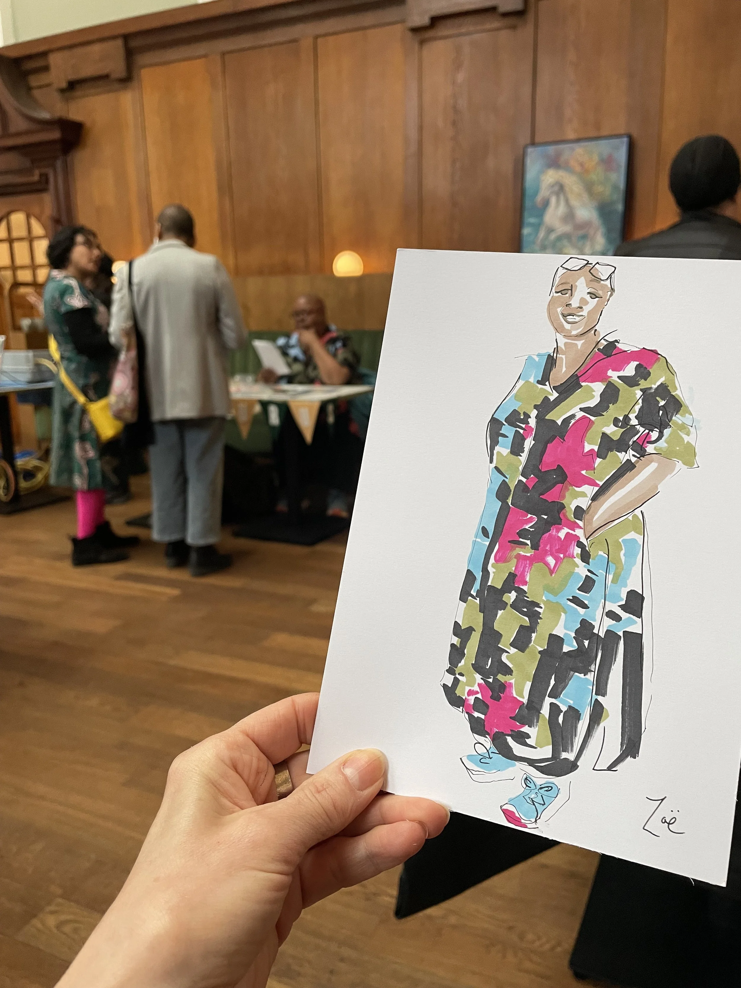 A person holding a colorful drawing of a woman with short hair, glasses on her head, and wearing a vibrant patterned outfit, in a room with wooden walls and artwork in the background.